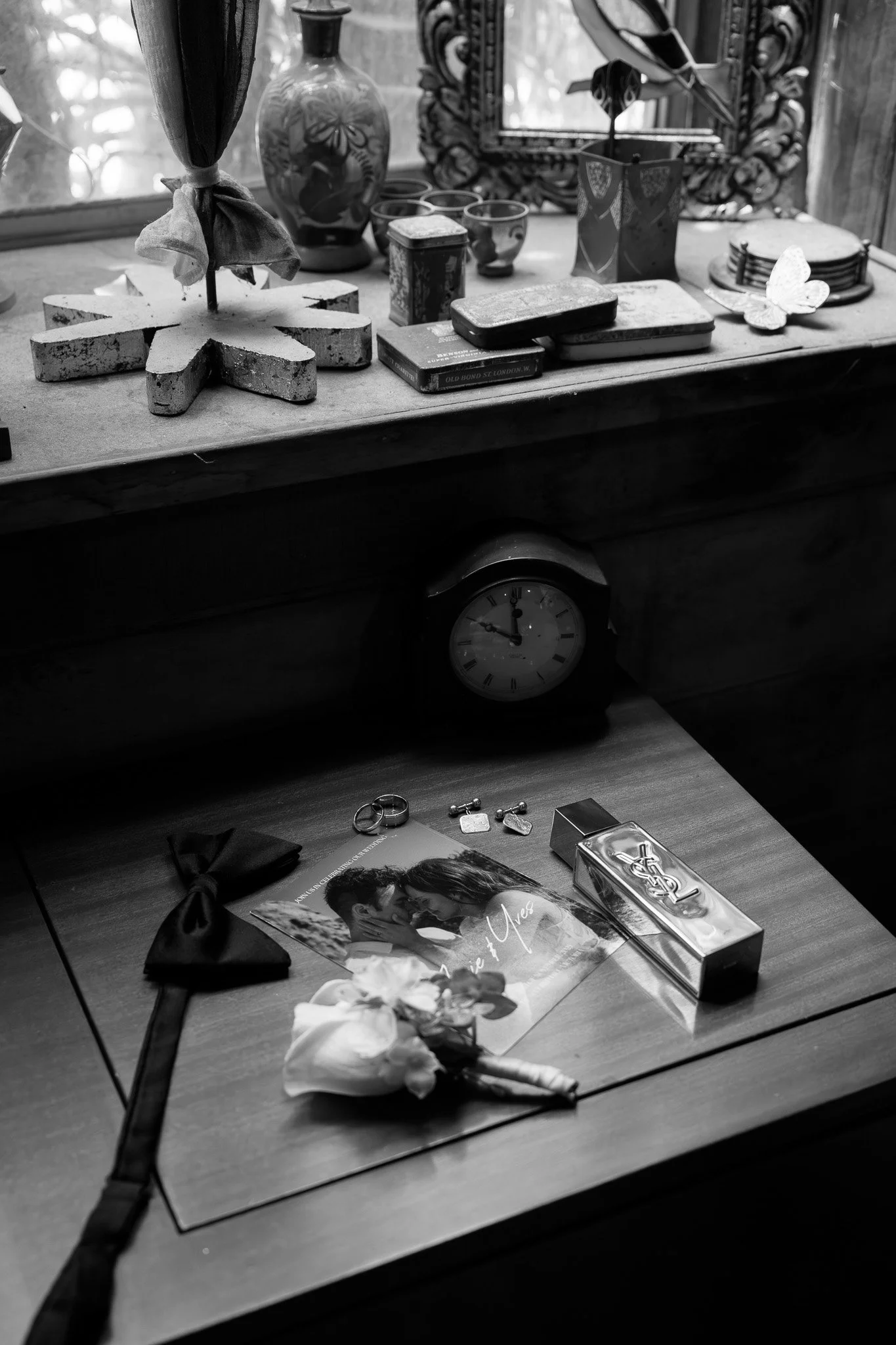 A wooden table with a wedding bouquet, a black bow tie, and a wedding photo of a couple. On the desk, there are wedding rings, keys, and a box of chocolates. In the background, a clock shows 1:11, and the top surface holds decorative items including 
