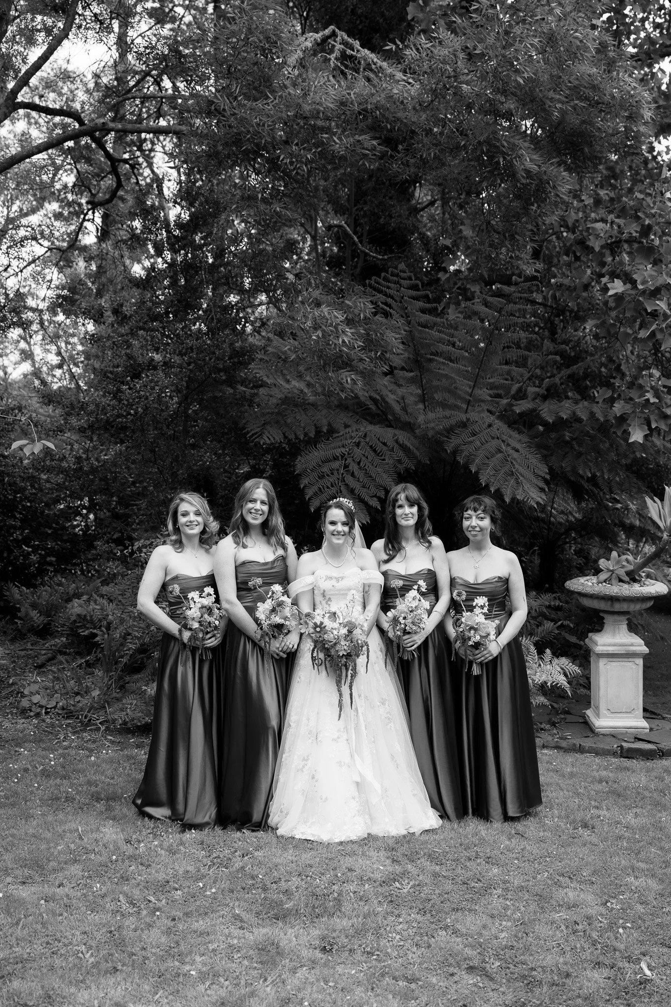 Black and white photo of a bride and four bridesmaids standing outdoors in front of trees and foliage, all smiling and holding bouquets.