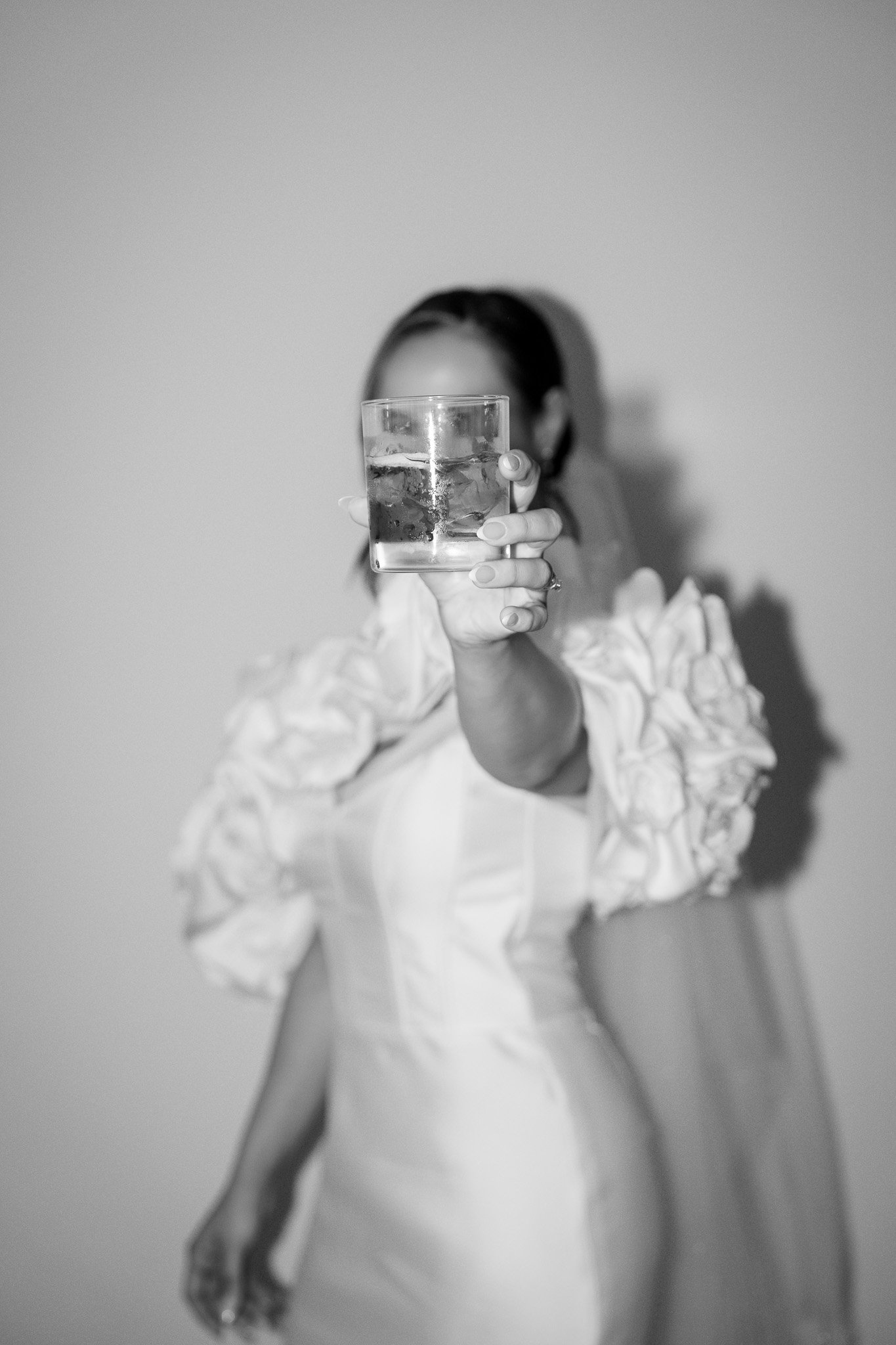 A woman in a white dress with ruffled sleeves holding a glass of water toward the camera, obscuring her face.