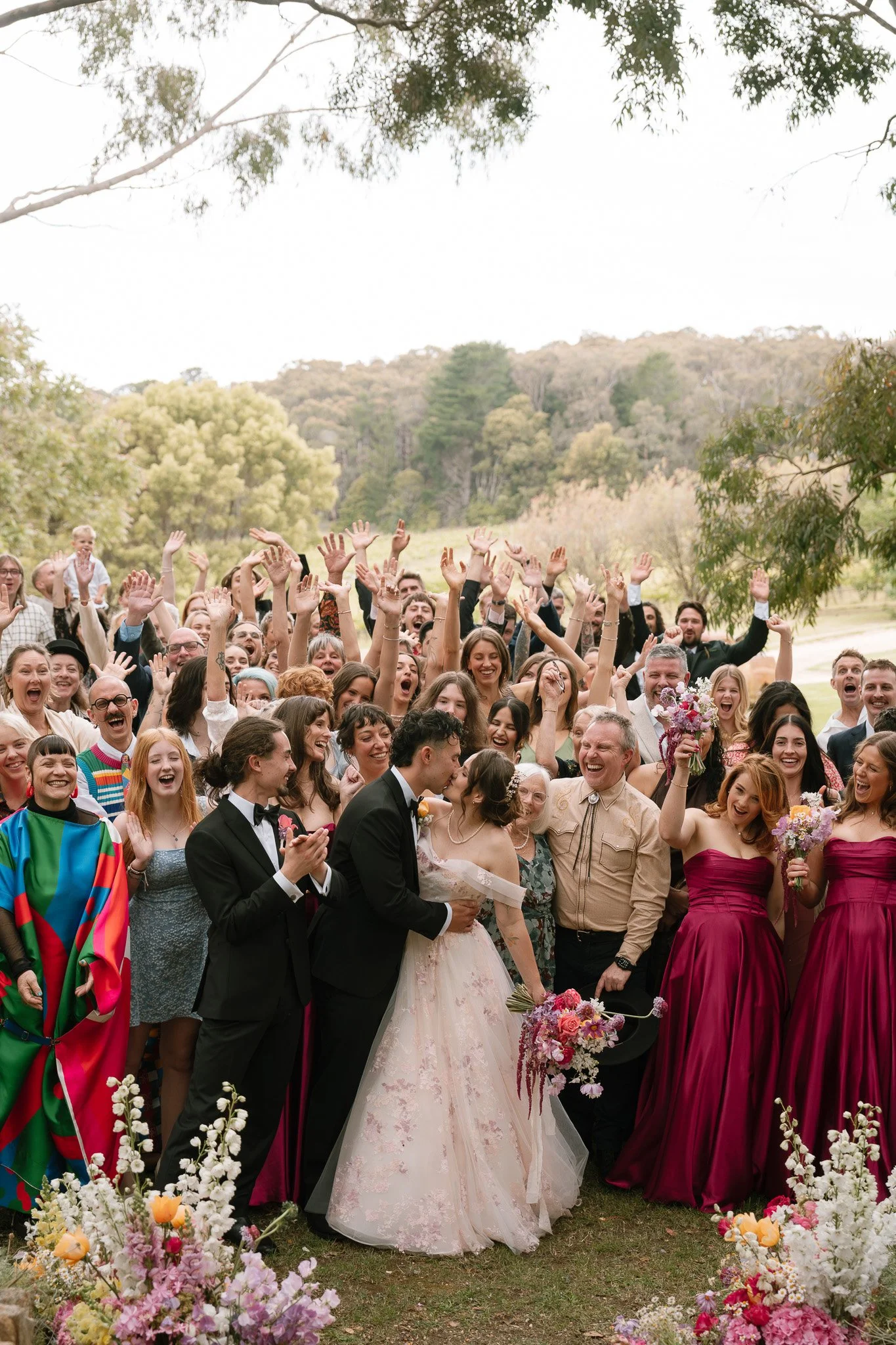 A bride and groom celebrating with a large group of wedding guests outdoors, surrounded by trees and flowers.