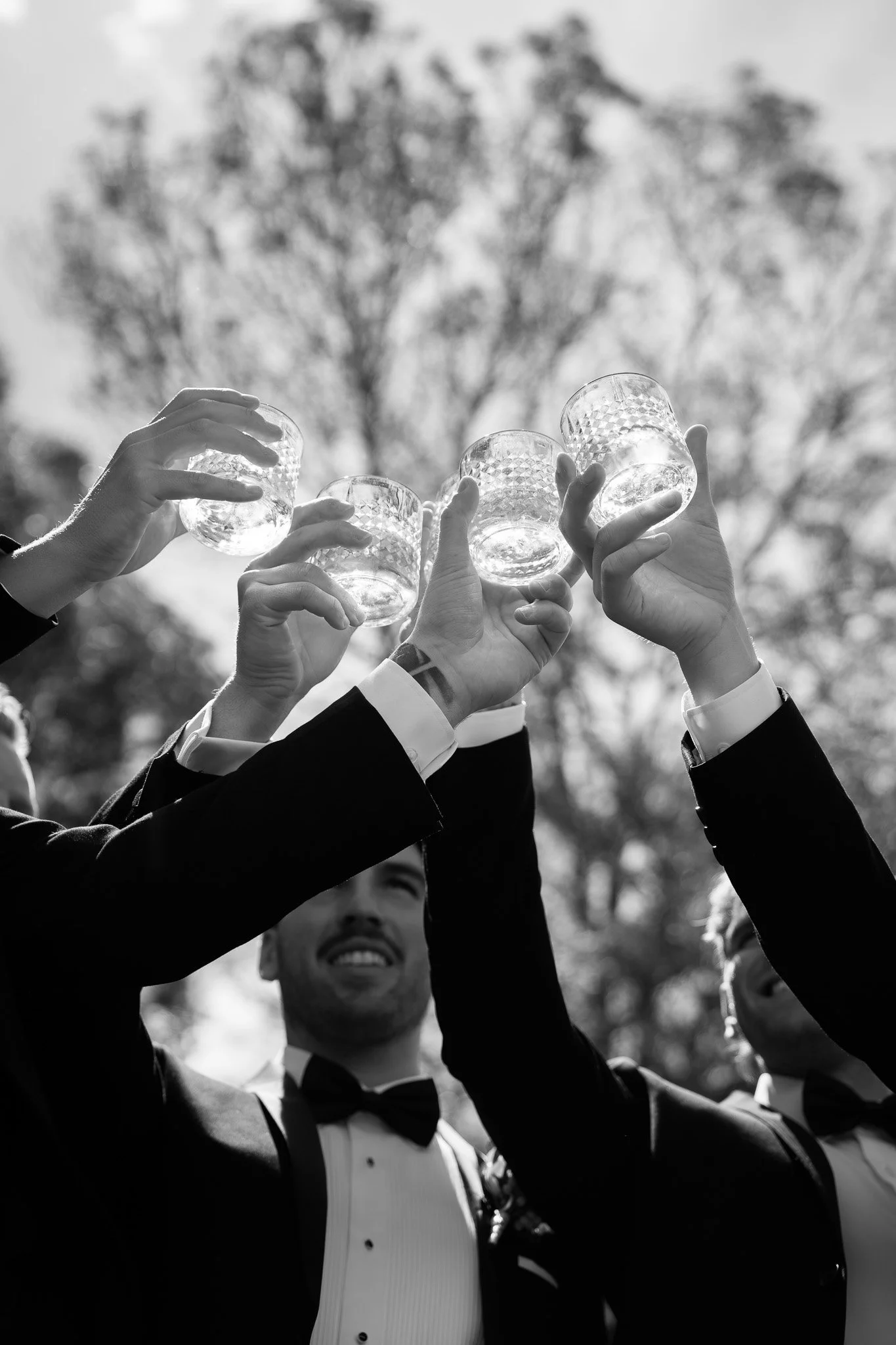 Group of people in tuxedos raising glasses outdoors in a toast, black and white photograph, trees in background.