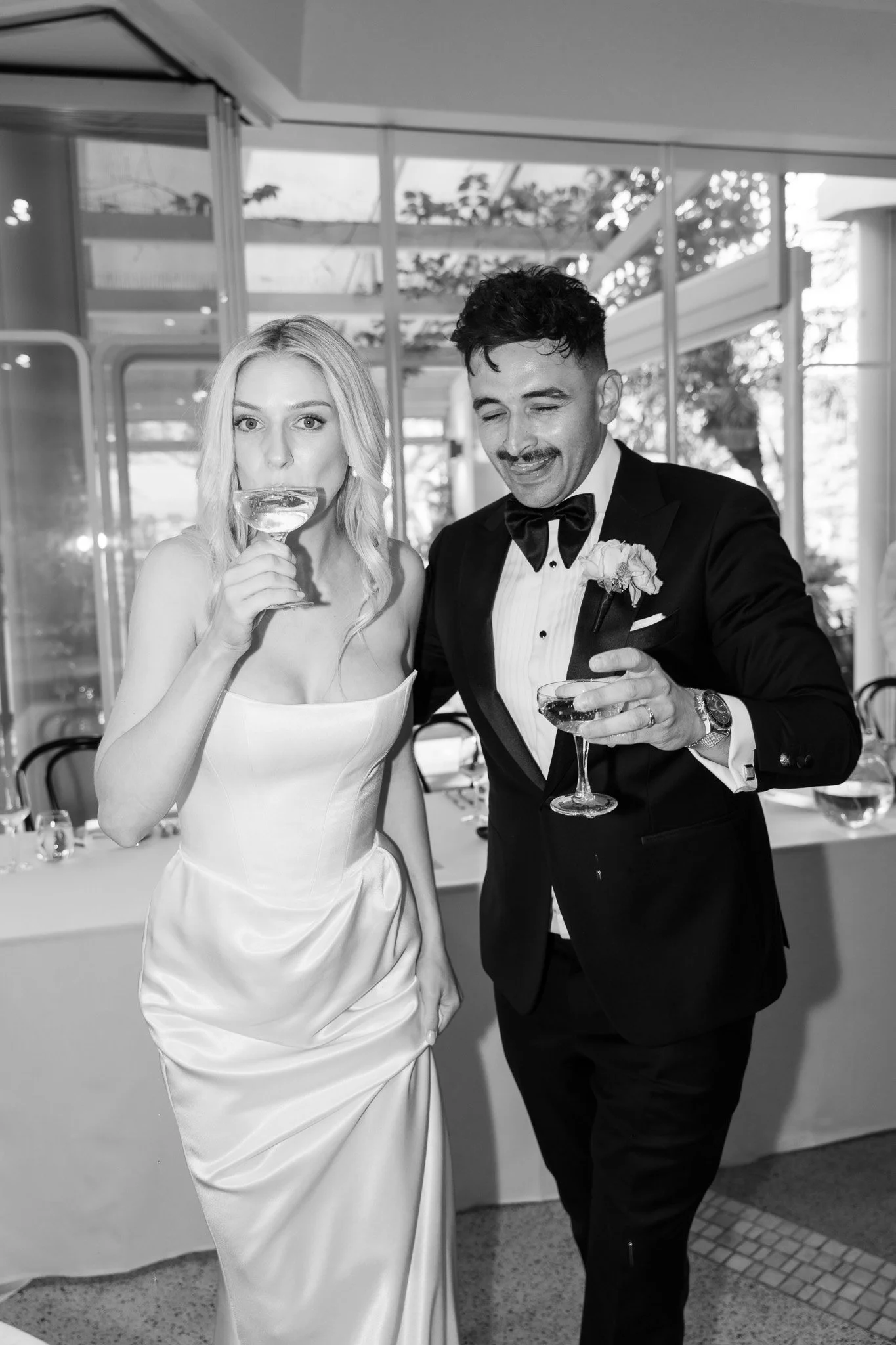 Black and white photo of a bride and groom holding cocktails, attending a wedding reception indoors.