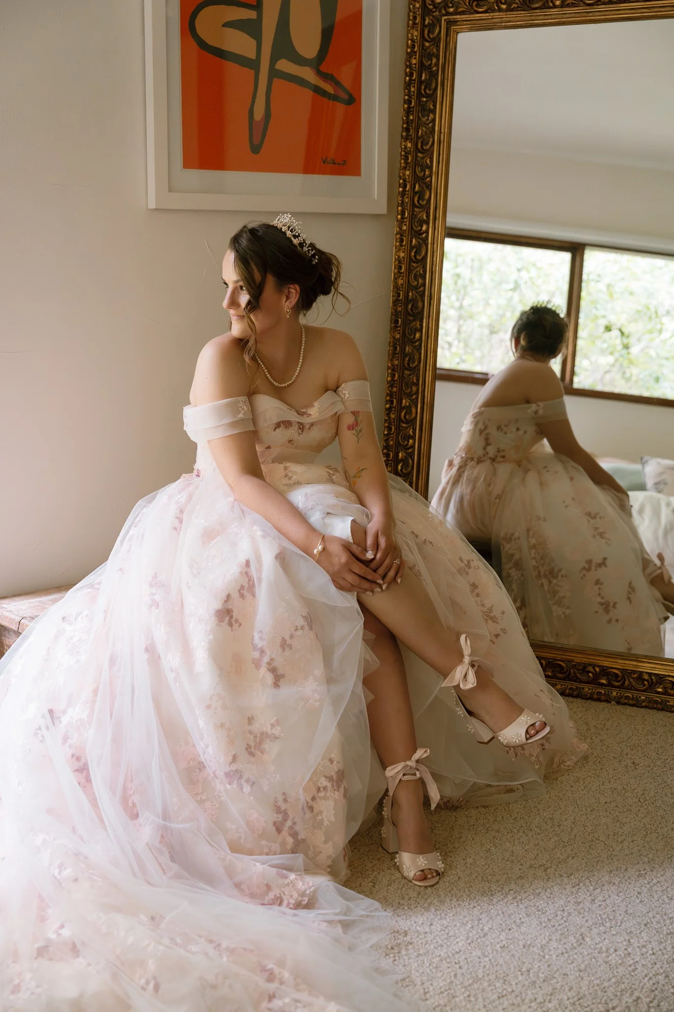 A bride in a wedding dress sitting on a bench, adjusting her shoe, with a large mirror reflecting her and a window showing outdoor greenery.
