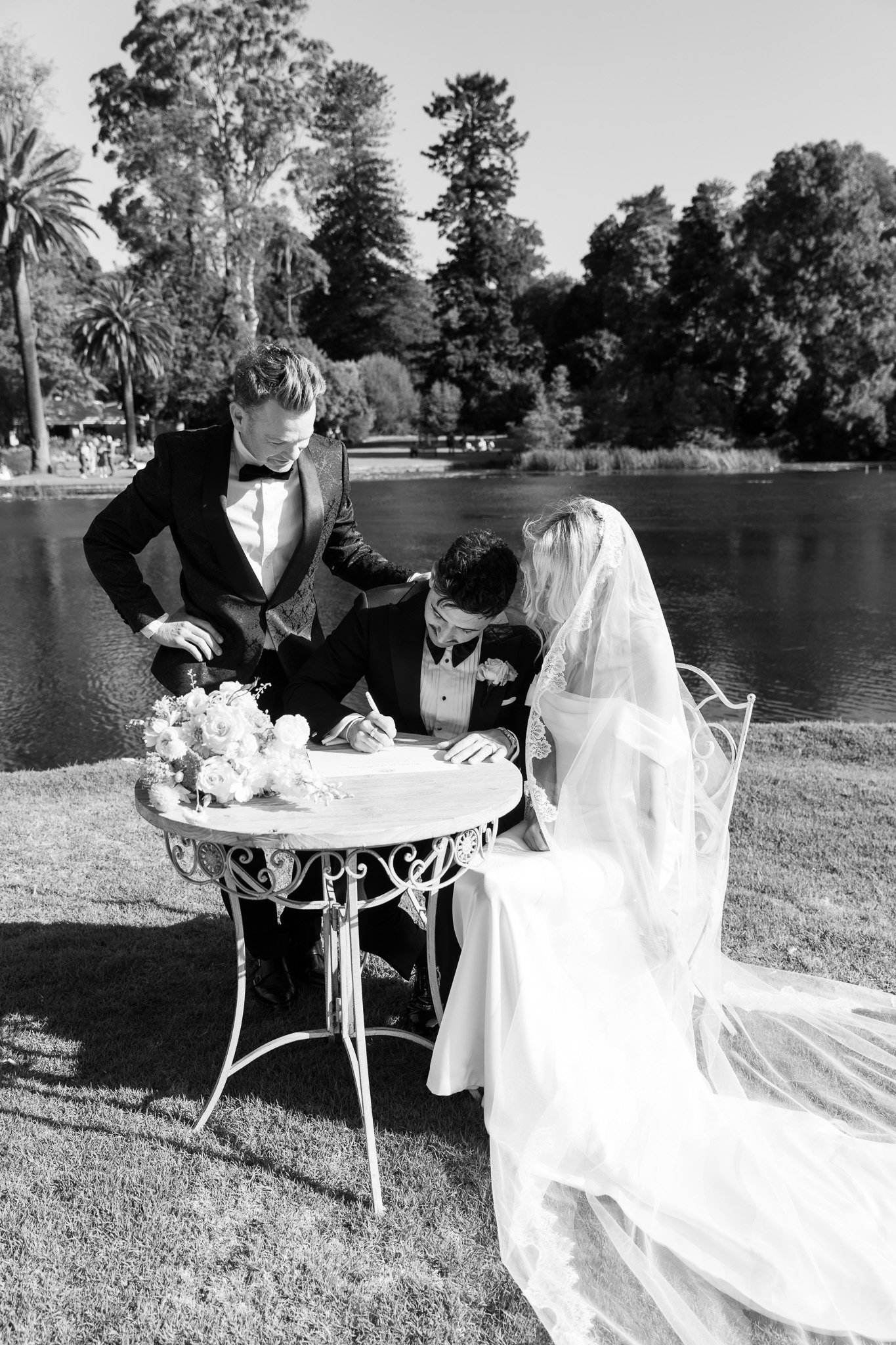 Black and white photo of a wedding ceremony outdoors by a lake. A bride and groom are seated at a small table, signing a document, with a man in a tuxedo standing beside them.