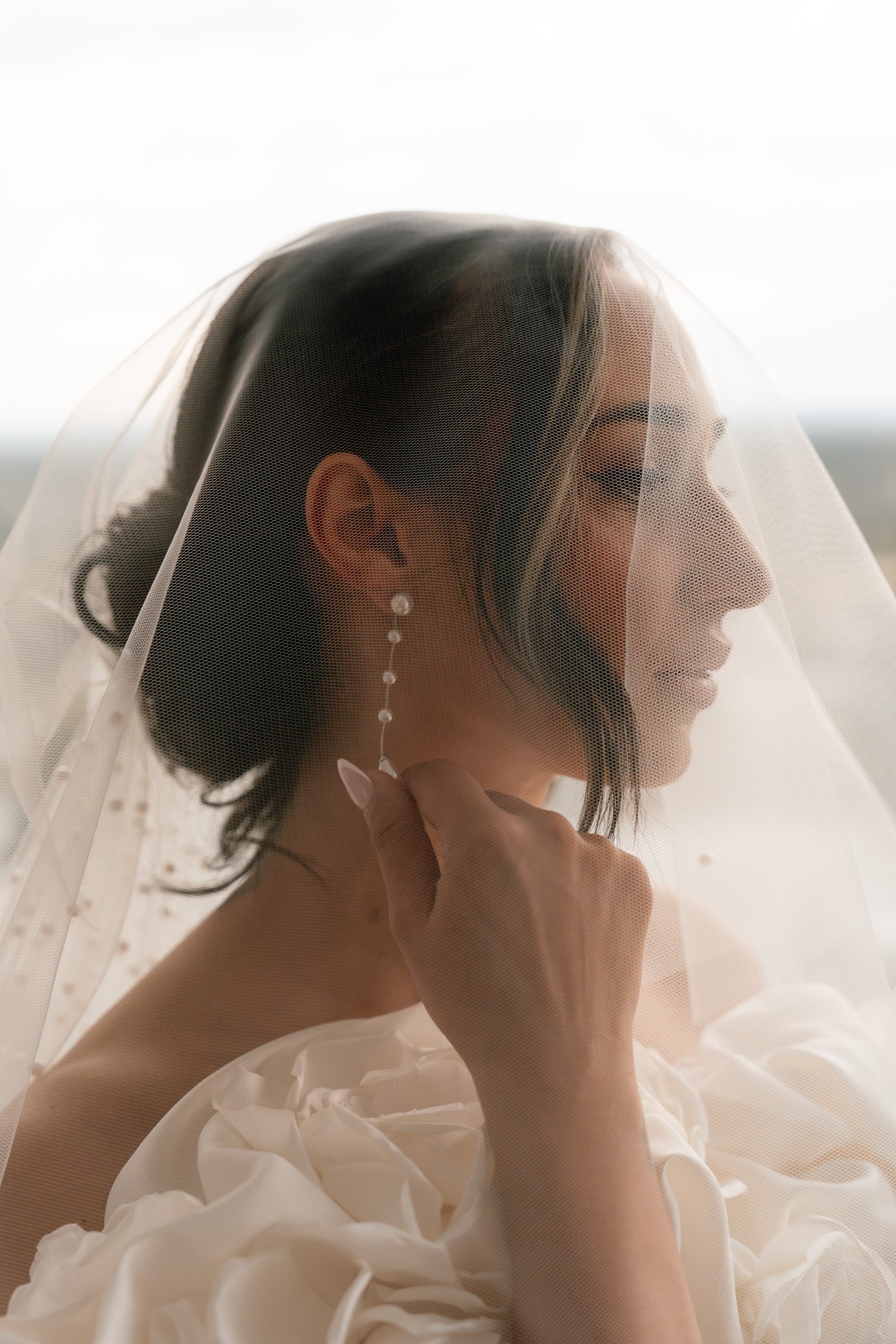 A woman in a wedding dress holding an earring with a veil over her face.