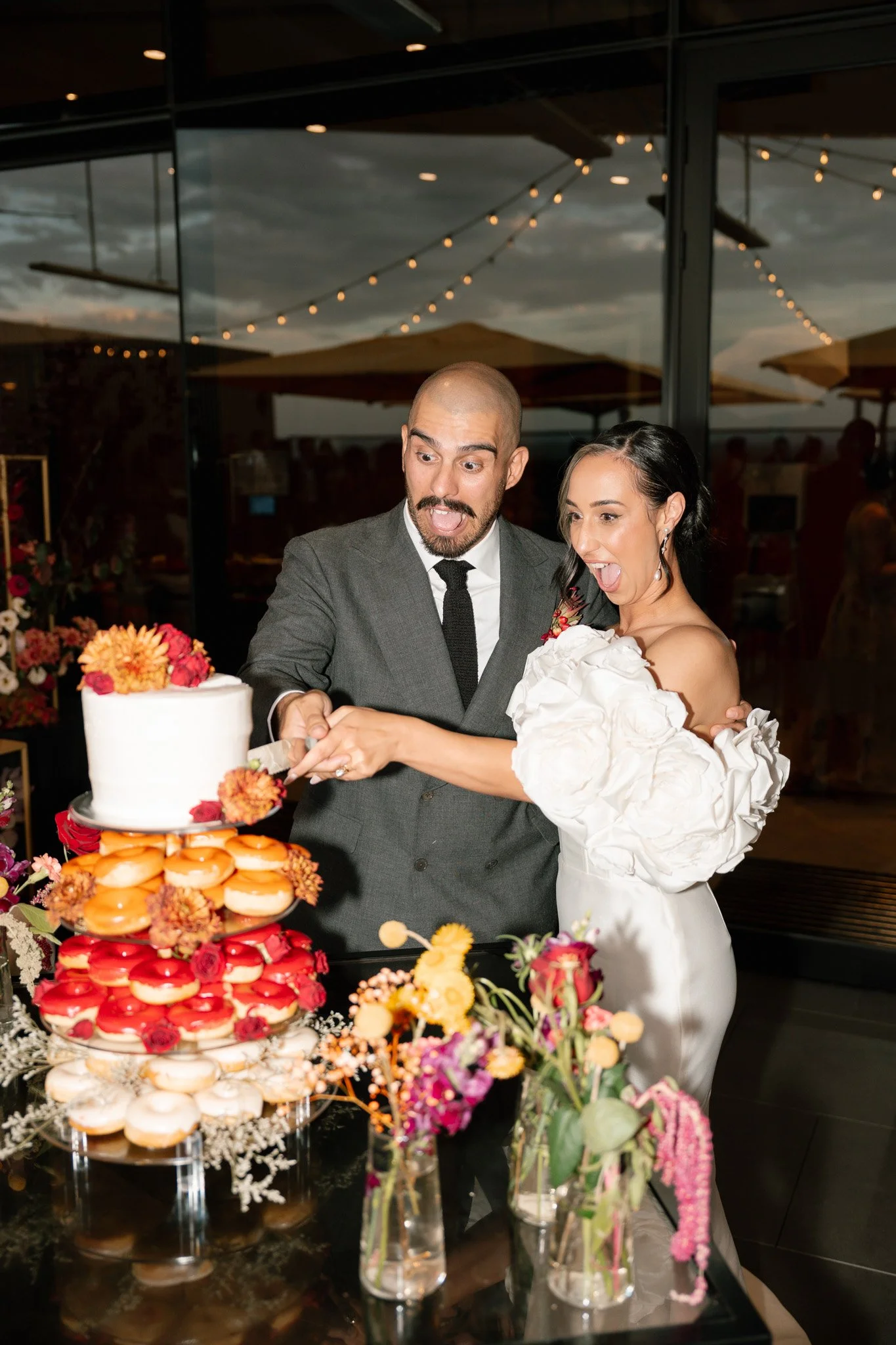 A couple at their wedding reception cut a wedding cake together, surrounded by colorful flowers and desserts, with string lights and windows showing an outdoor scene in the background.