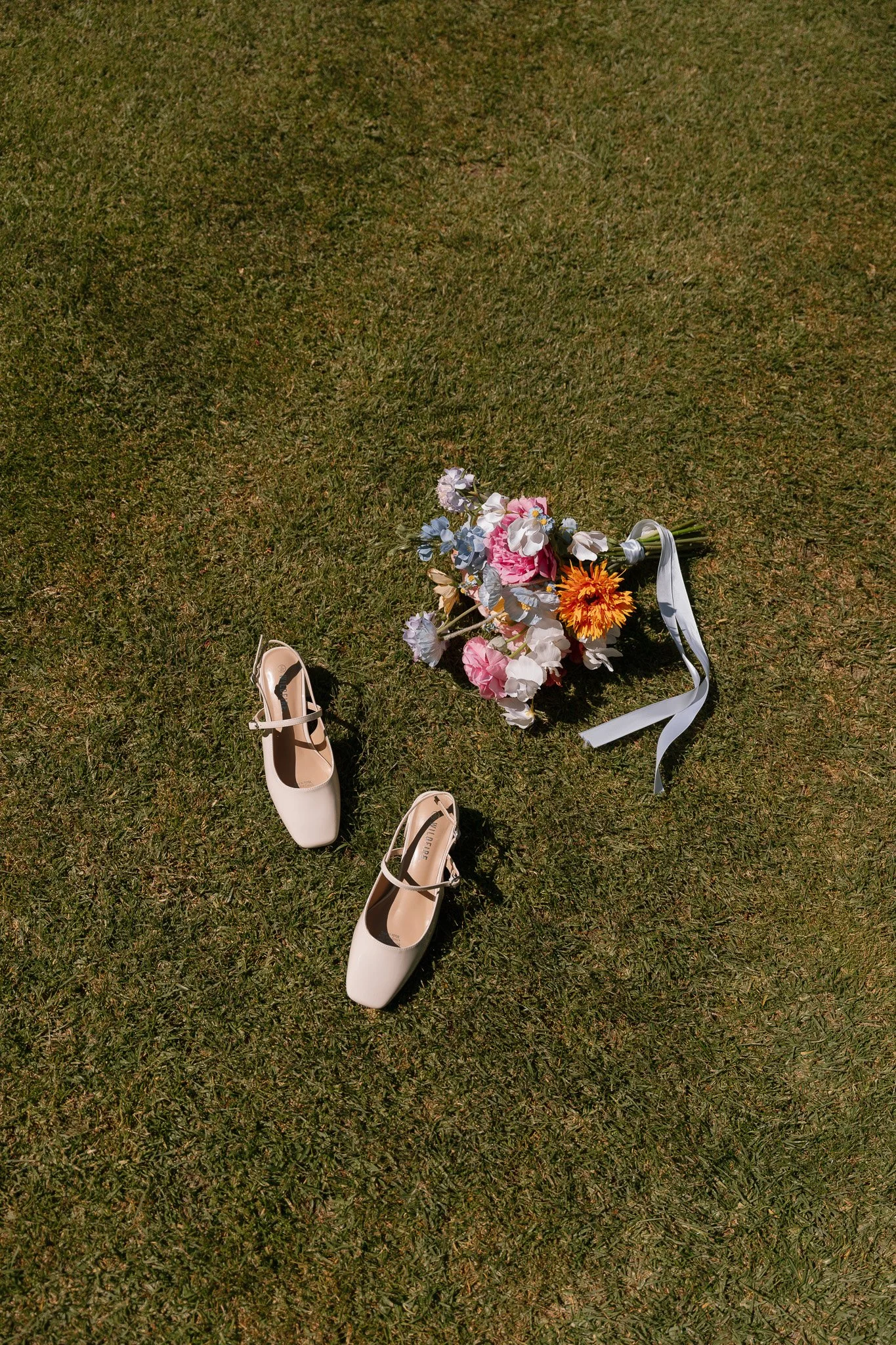 A pair of beige high-heeled shoes and a colorful bouquet of flowers laid on a grassy surface.