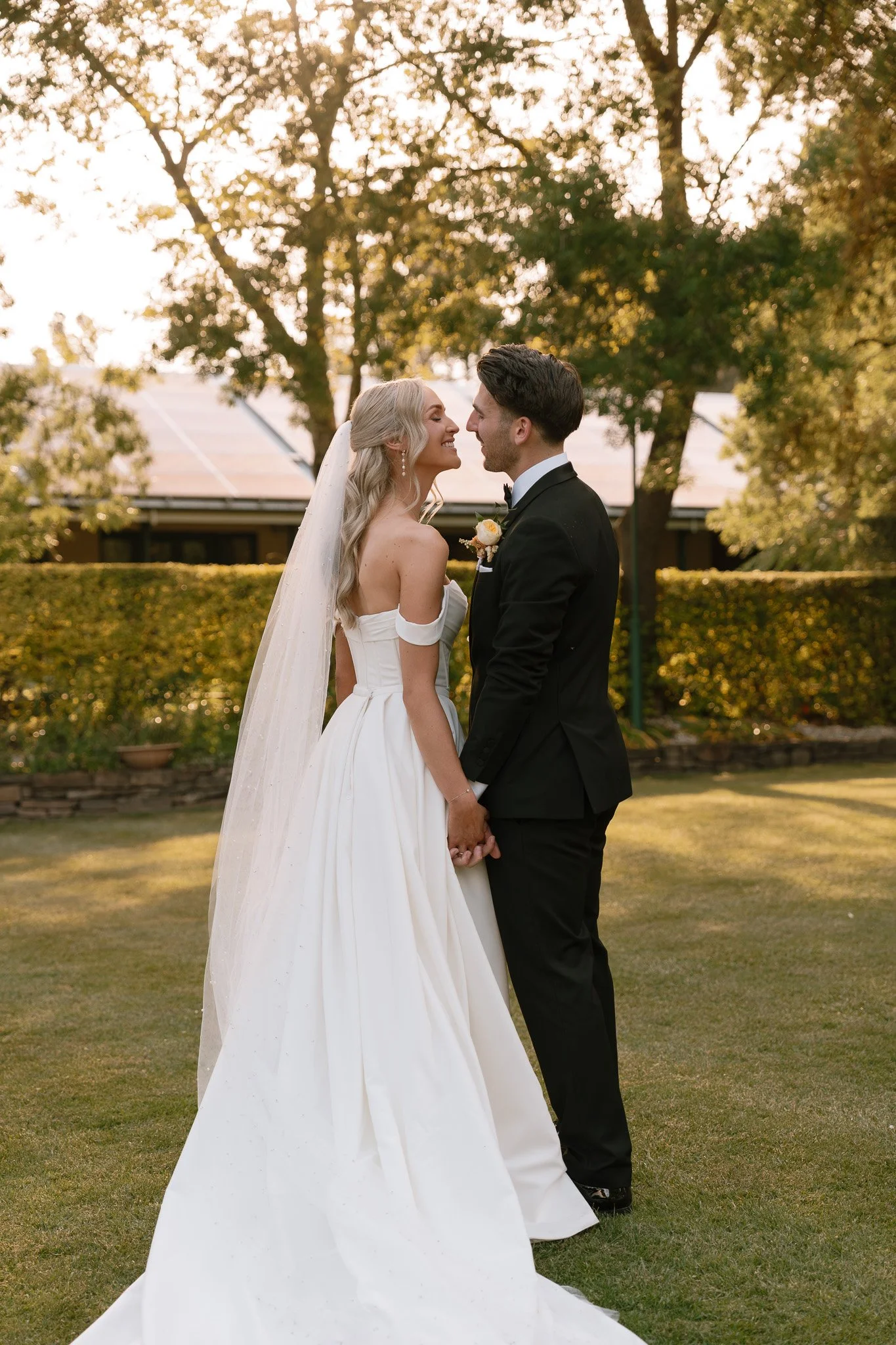 A bride and groom face each other and hold hands in a grassy outdoor area during sunset, surrounded by trees and hedges.