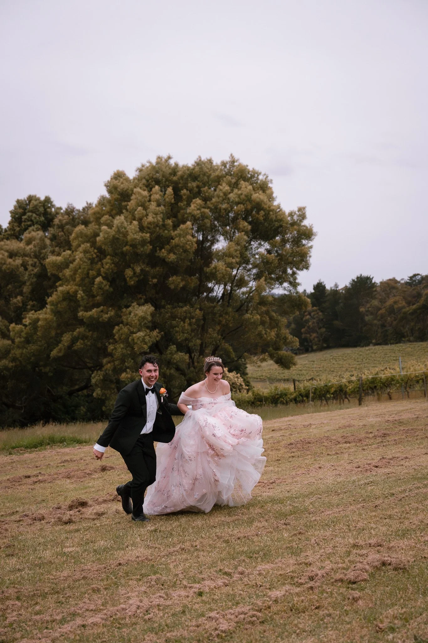 A bride and groom running together outdoors on a grassy field with large trees and cloudy sky in the background.