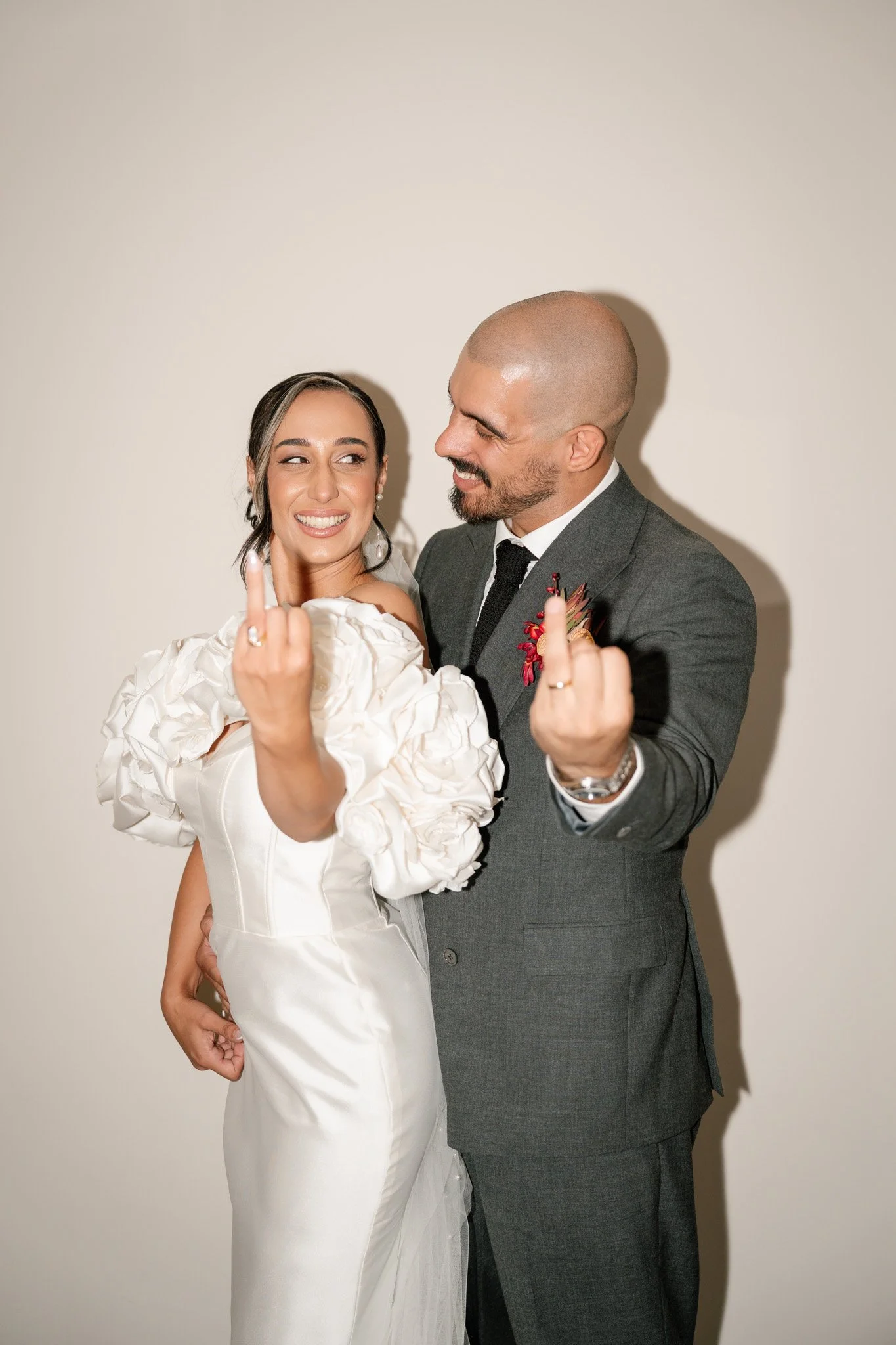 A newlywed couple making rude gestures, smiling, and looking at each other, against a plain beige background.
