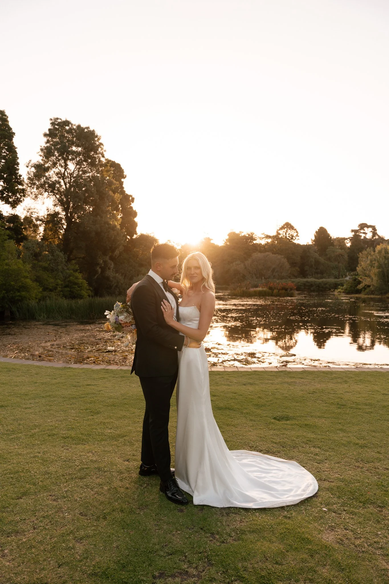 A bride and groom standing close together on a grassy area near a pond at sunset, with the sun setting behind trees in the background.
