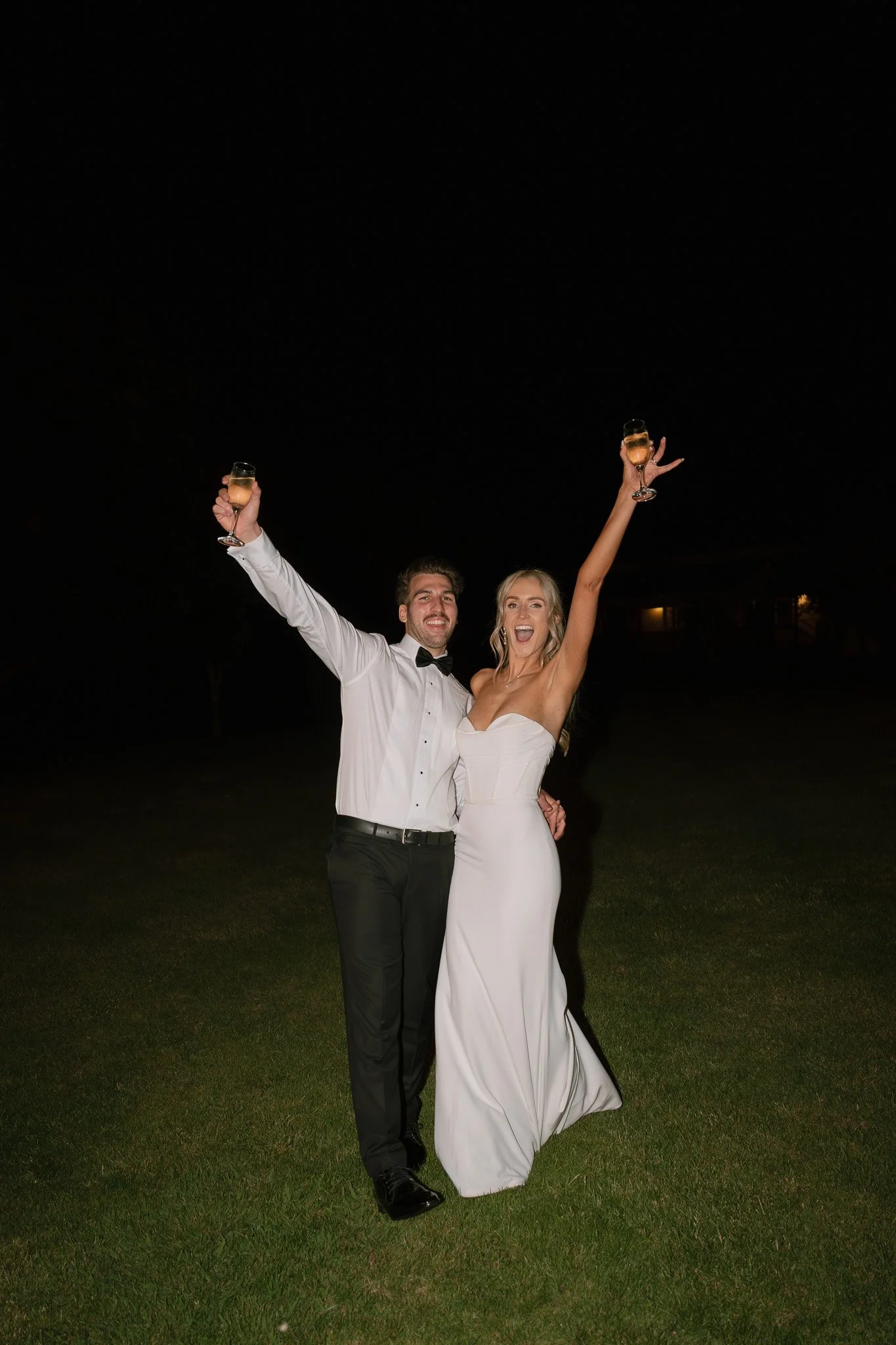 A newlywed couple celebrating at night, holding champagne glasses up, standing on grass, with a dark background.