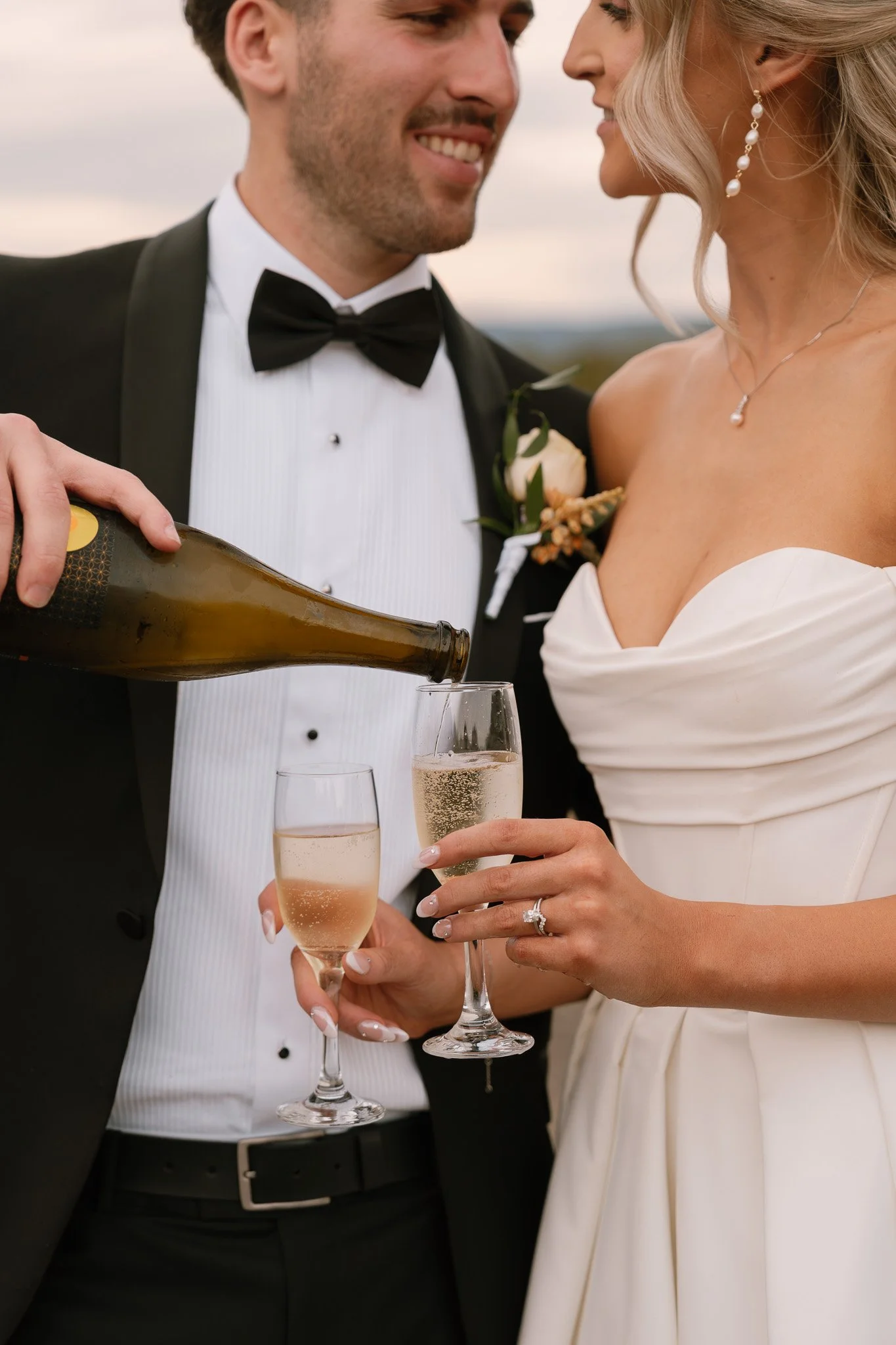A newlywed couple in formal wedding attire sharing a toast with champagne. The groom in a tuxedo is pouring champagne into the bride's glass, and they are smiling at each other.