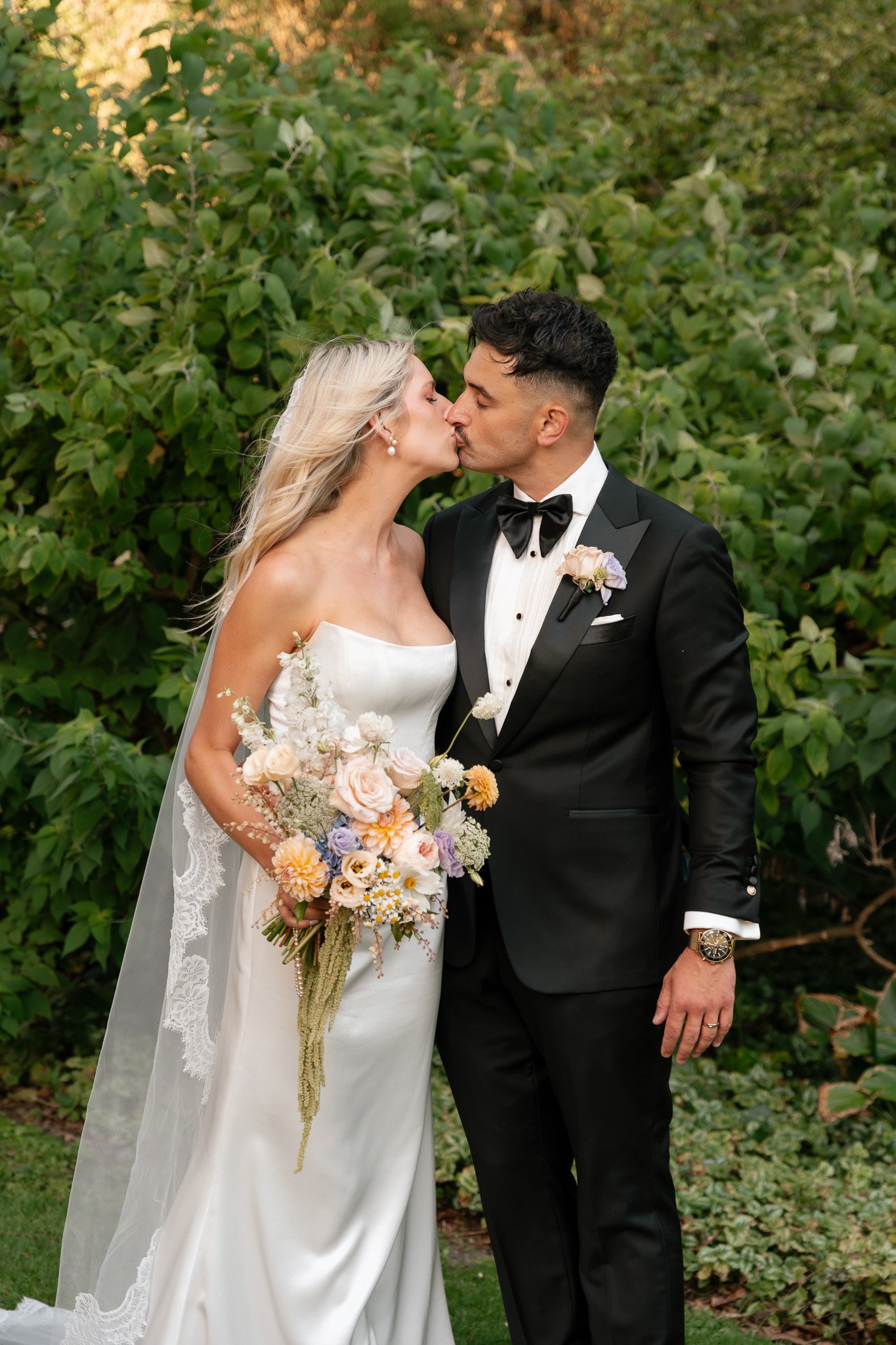 A newlywed couple kissing outdoors, the bride holding a colorful bouquet, the groom in a black tuxedo with a bow tie, surrounded by green foliage.