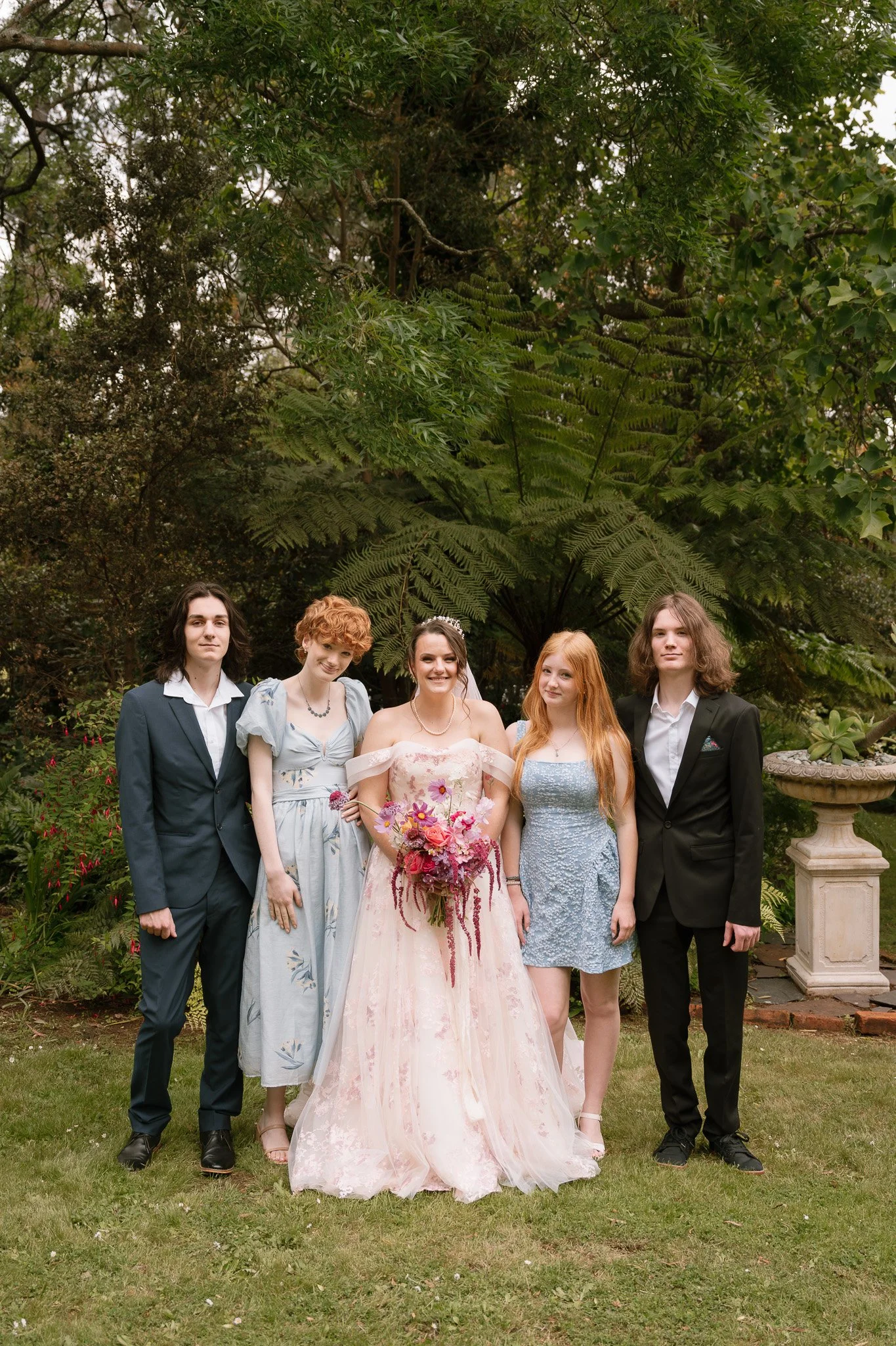 Group of five young people dressed in formal attire standing outdoors in front of lush greenery and trees, with a decorative stone planter on the right.