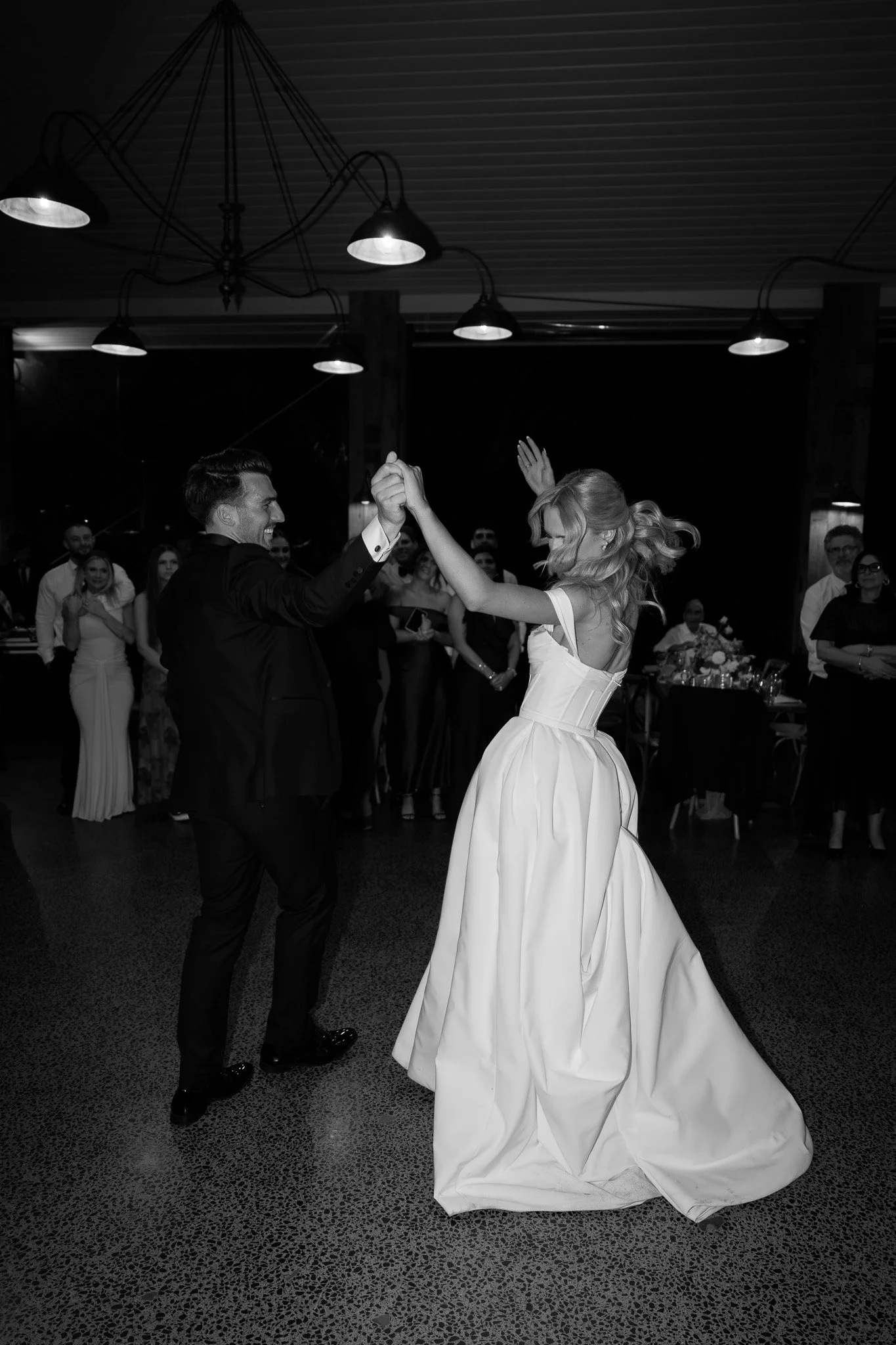 Black and white photo of a bride and groom dancing at their wedding reception, surrounded by guests.