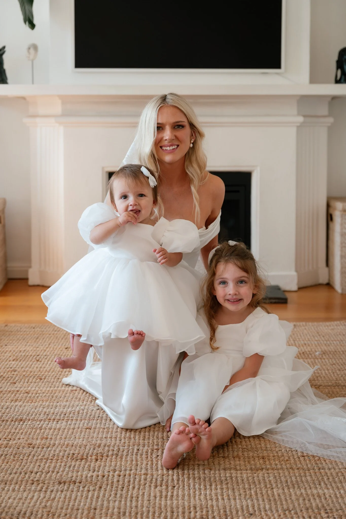 A blonde woman in a white dress with two young girls in white dresses, indoors in front of a fireplace, posing for a photo, all smiling and barefoot on a woven rug.