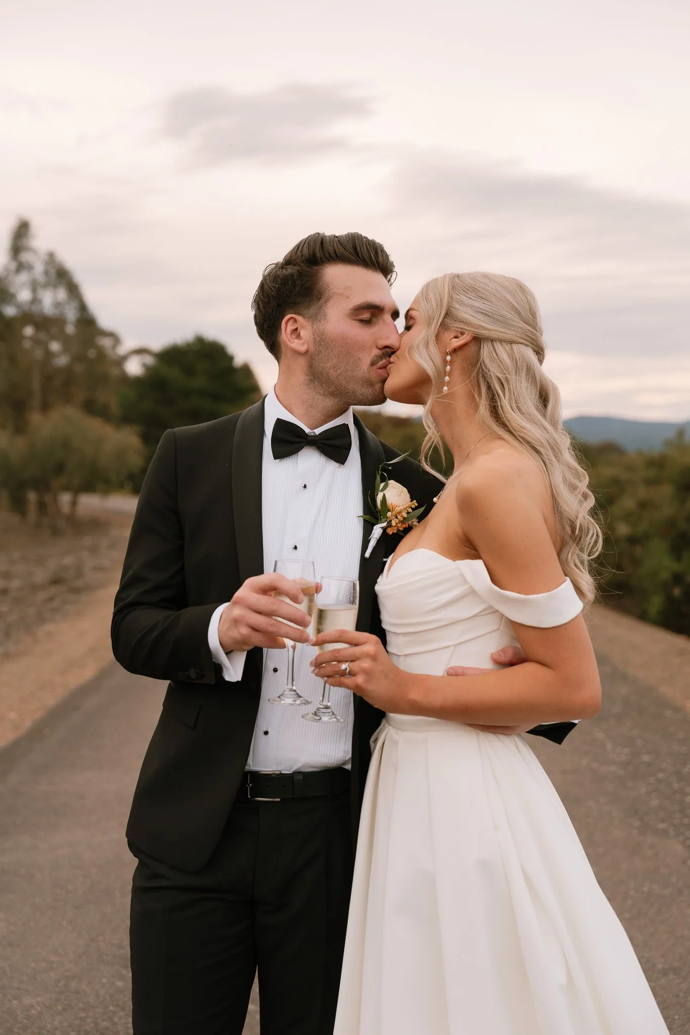 A newlywed couple, dressed in wedding attire, sharing a kiss while holding champagne glasses outdoors during daytime.