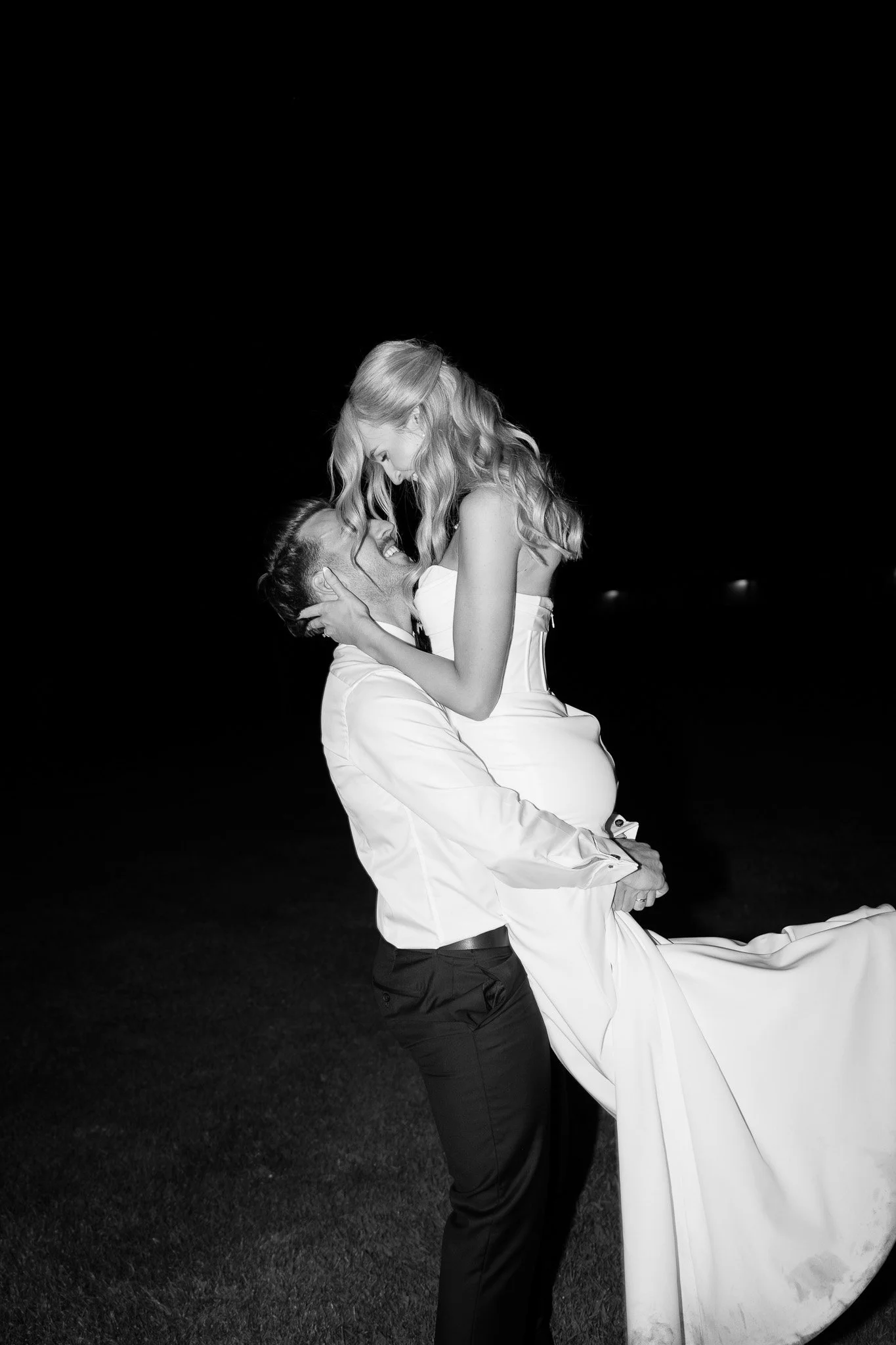 A couple on their wedding day, with the groom lifting the bride and both smiling, in black and white.