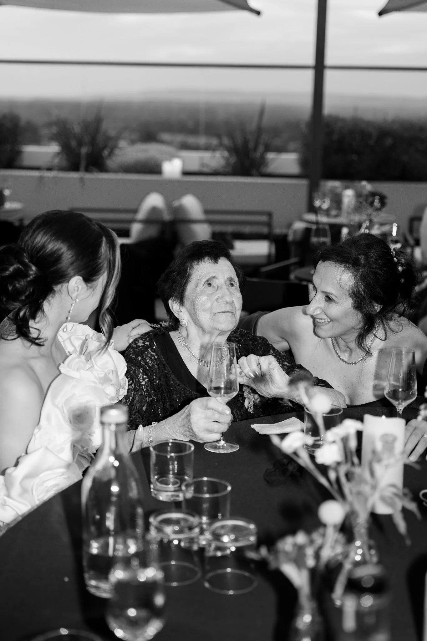 Three women at a dinner table, one elderly woman with a glass of wine, smiling and engaging with two younger women, one on each side, in a celebration setting.