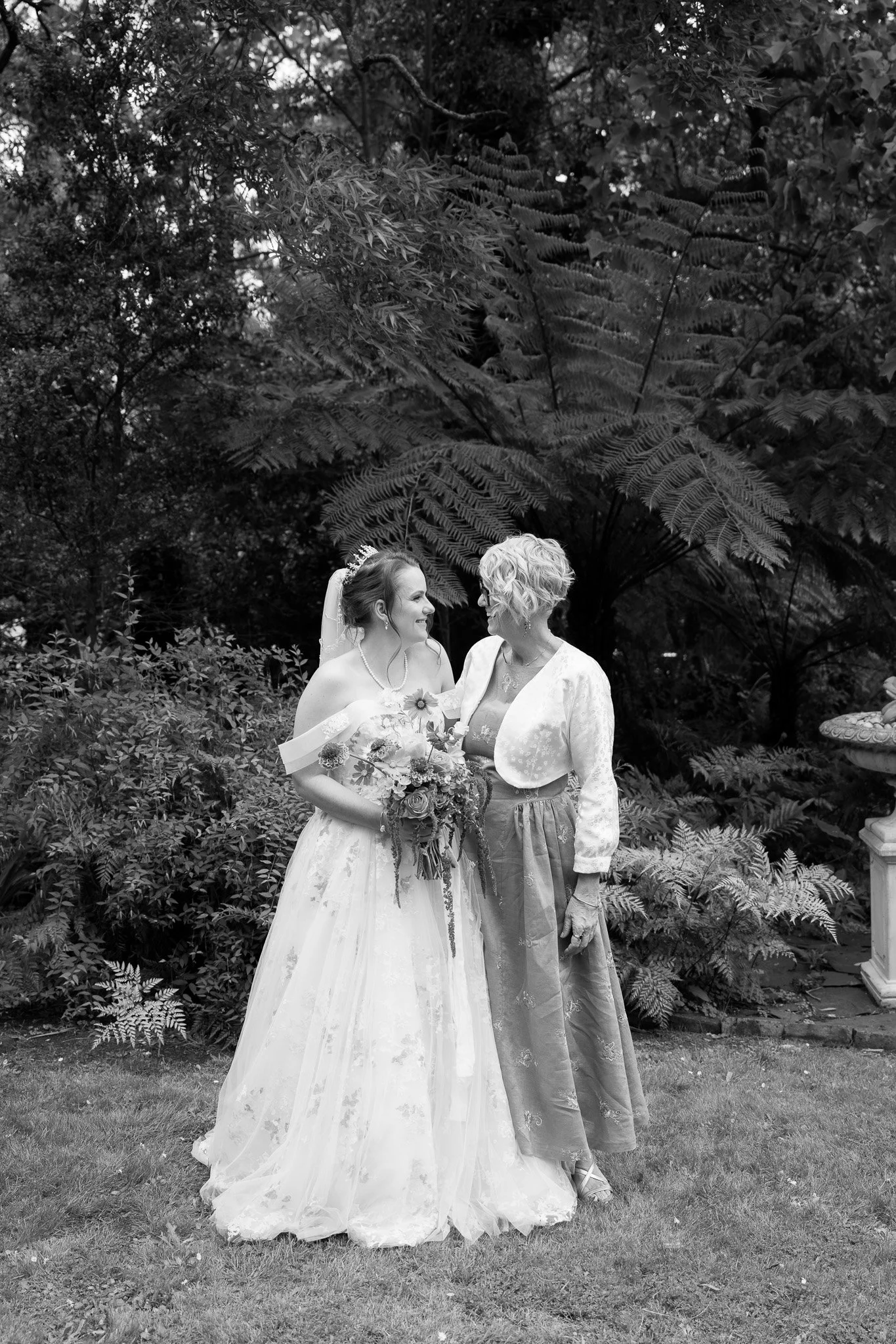 A bride holding a bouquet and a woman, likely her mother, standing outdoors in front of lush foliage, sharing a joyful moment.