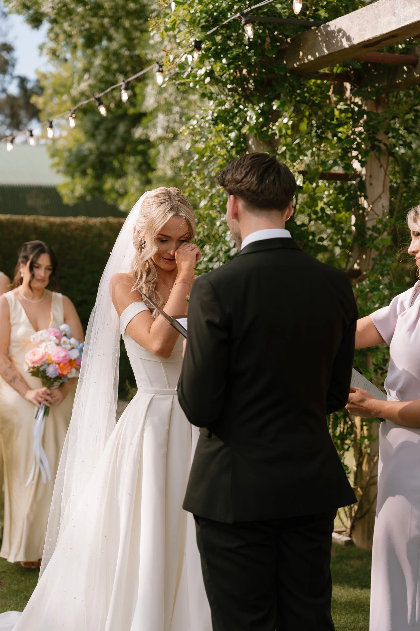 A bride and groom exchanging vows during a wedding ceremony outdoors with bridesmaids and greenery in the background.