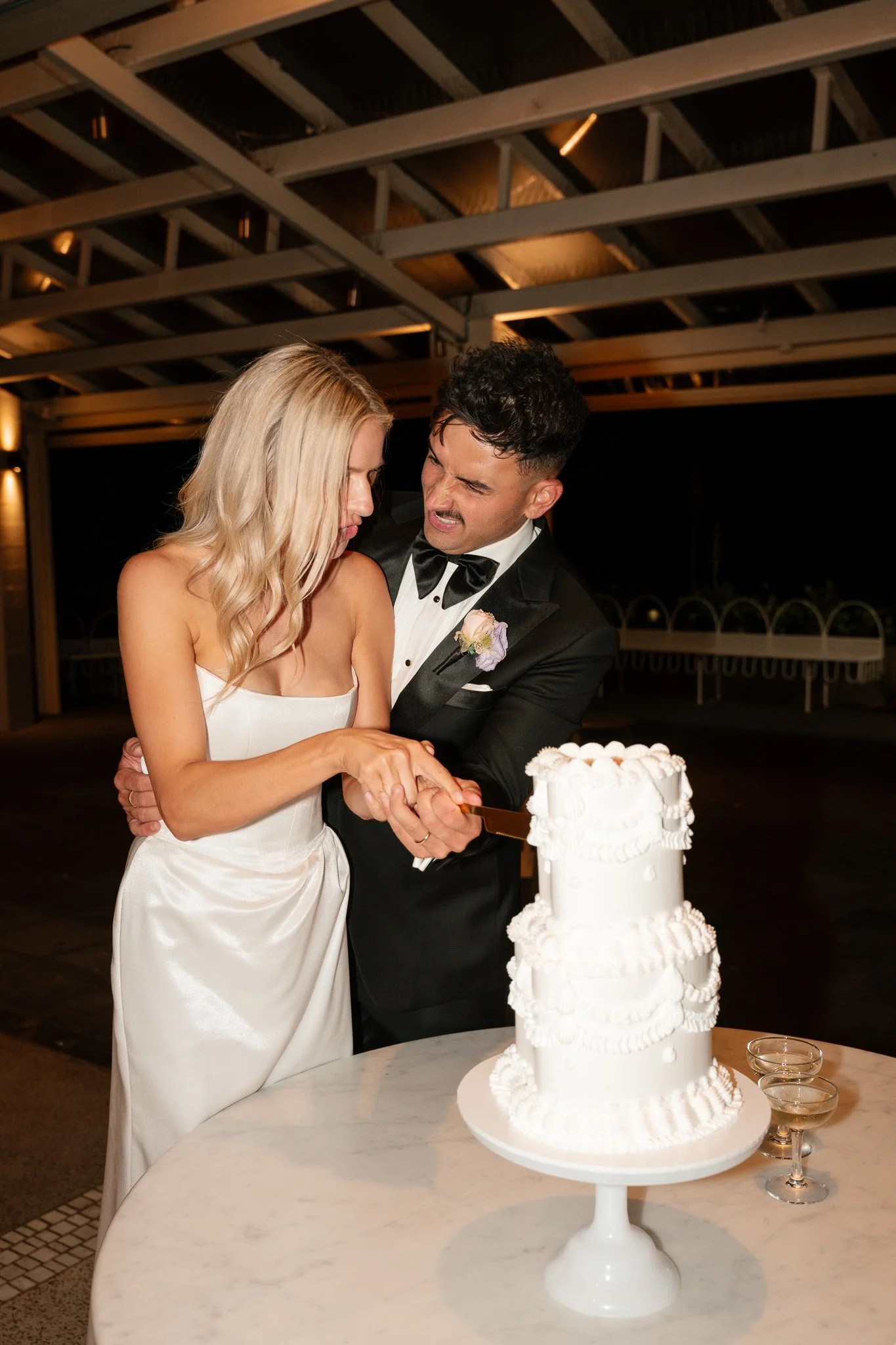 A bride and groom in formal wedding attire are cutting a white wedding cake together at a reception.