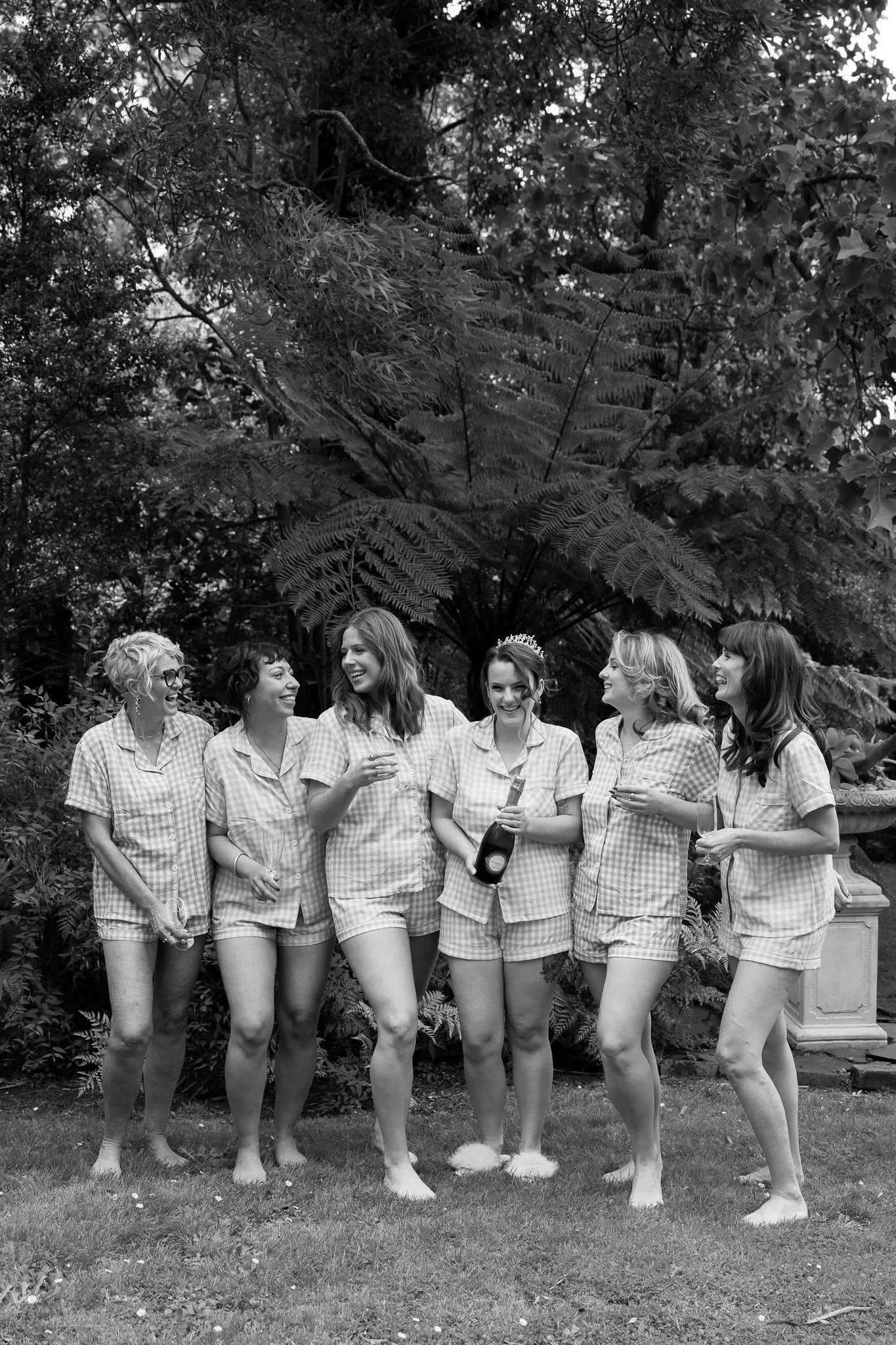 Group of women in pajamas celebrating with drinks outdoors, one wearing a tiara, greenery in the background.