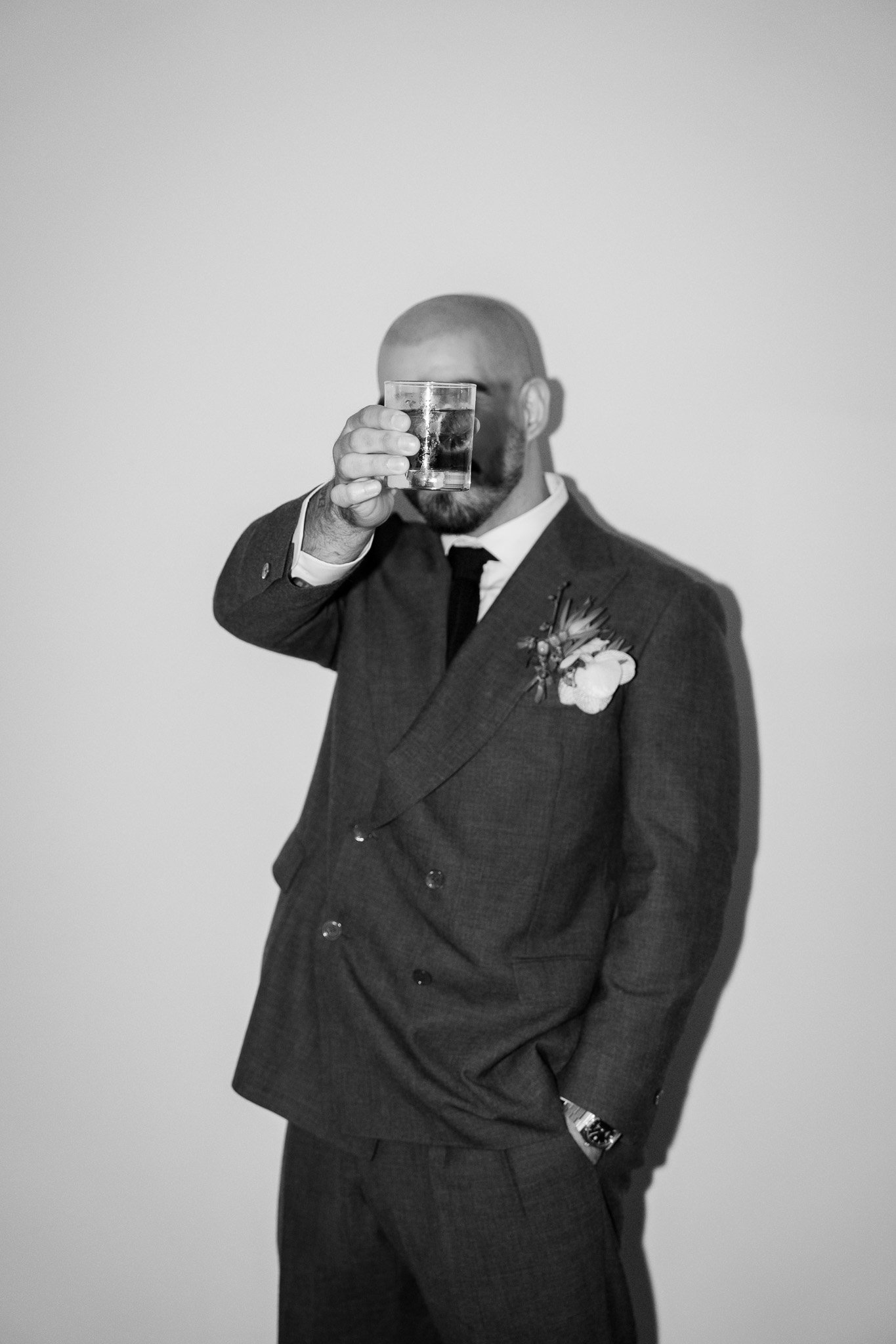A man in a suit with a boutonniere holding a glass of drink in front of his face, posing against a plain wall.