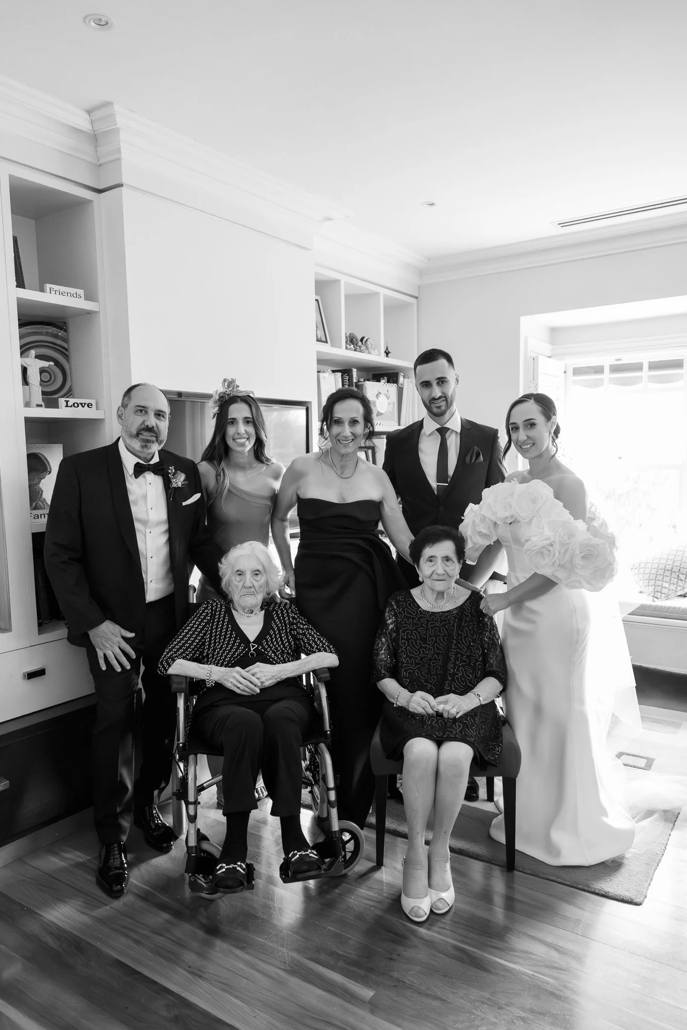 Family portrait at a wedding with six adults and two elderly women, some seated in wheelchairs, in a well-lit living room.