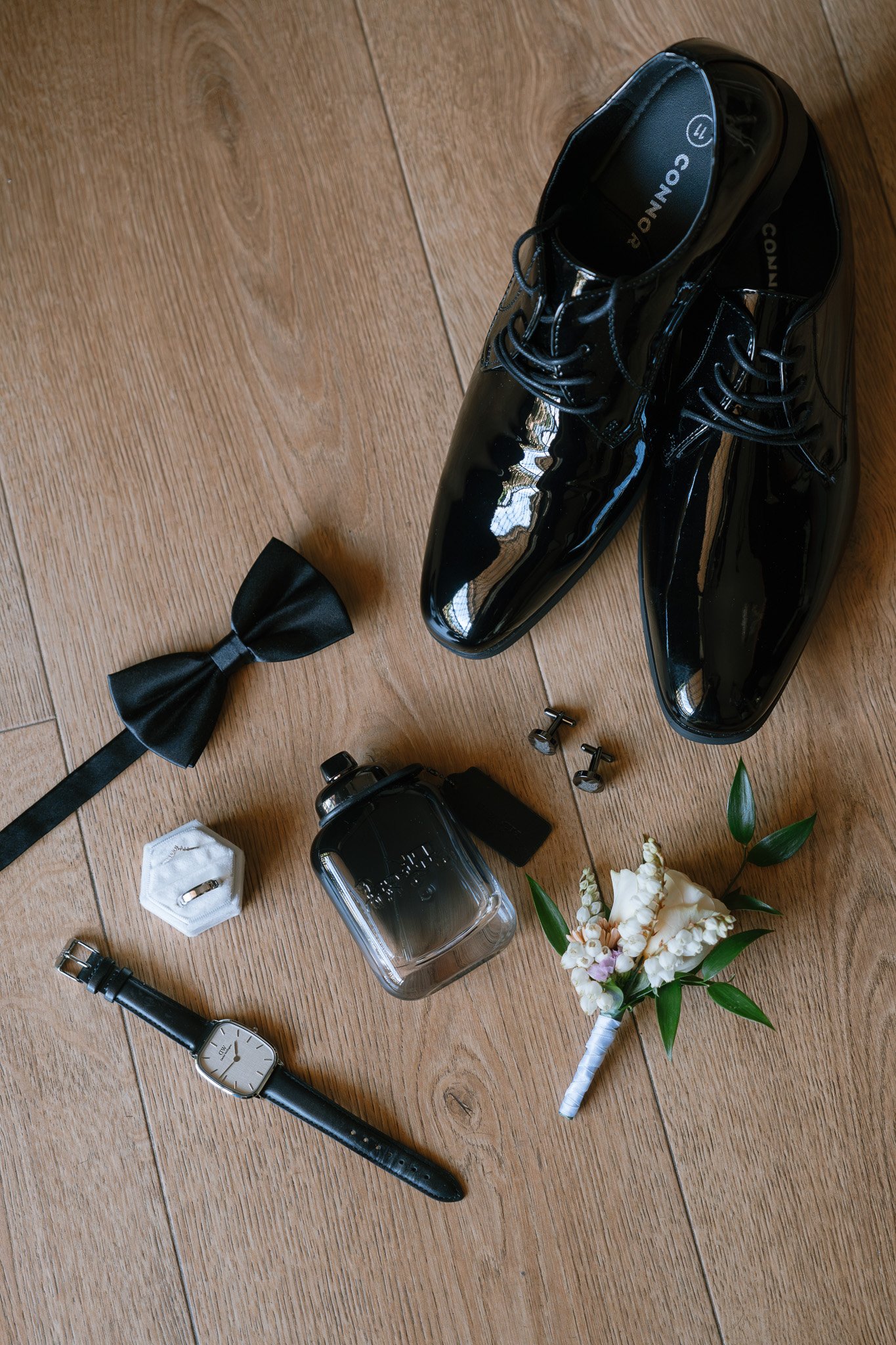 Wedding accessories including black shiny dress shoes, black bow tie, men's wristwatch, perfume bottle, wedding ring in a box, cufflinks, boutonniere, and a small glass bottle, all arranged on a wooden surface.