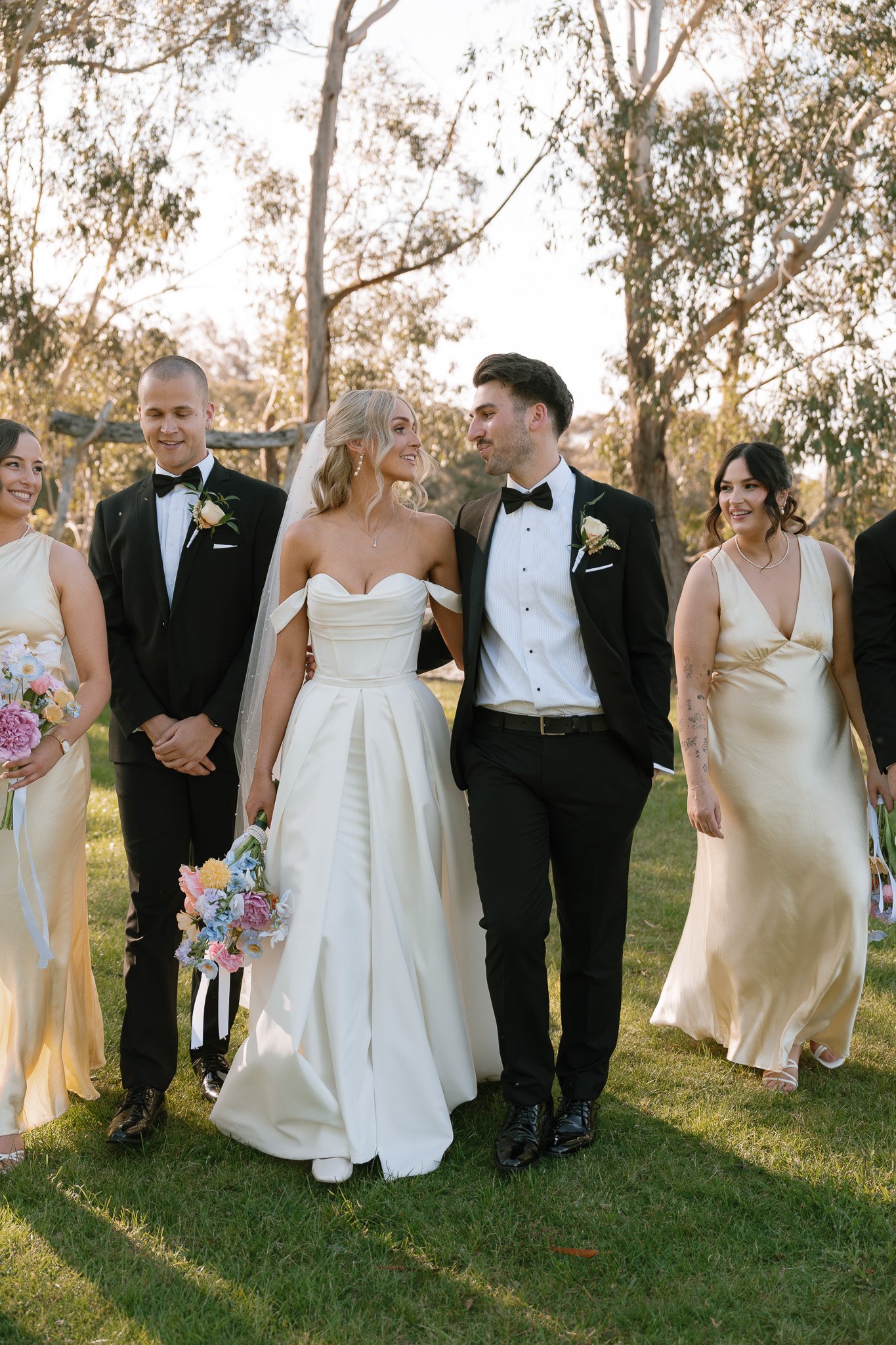 A group of wedding party members, including a bride and groom, walking outdoors on grass with trees and sunlight in the background. The bride is wearing a strapless white wedding dress, and the groom is in a black tuxedo with a bow tie. They are smil