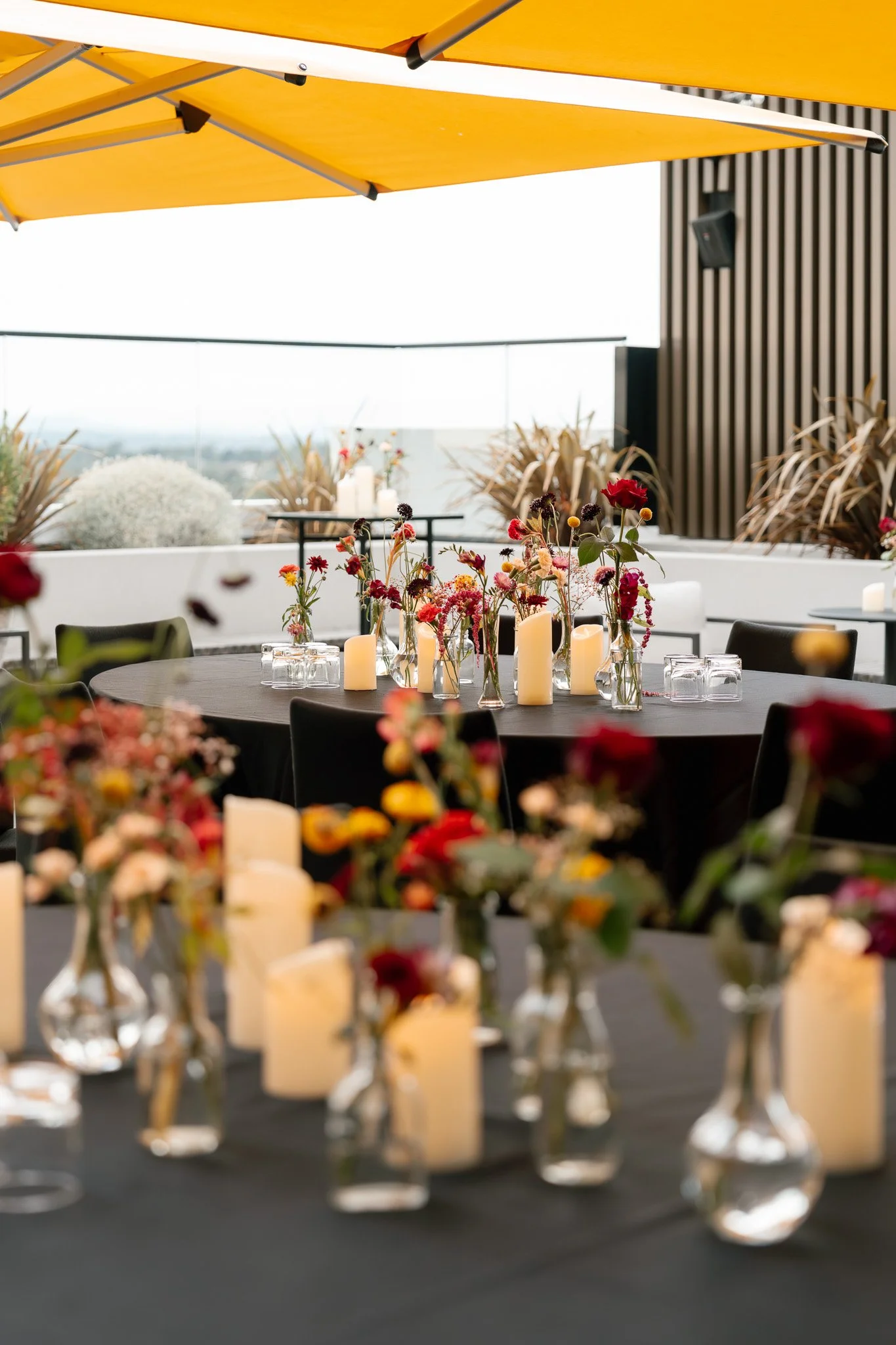 A decorated outdoor dining area with black tables, tulip-shaped vases filled with colorful flowers, and electric candles, under a yellow umbrella.