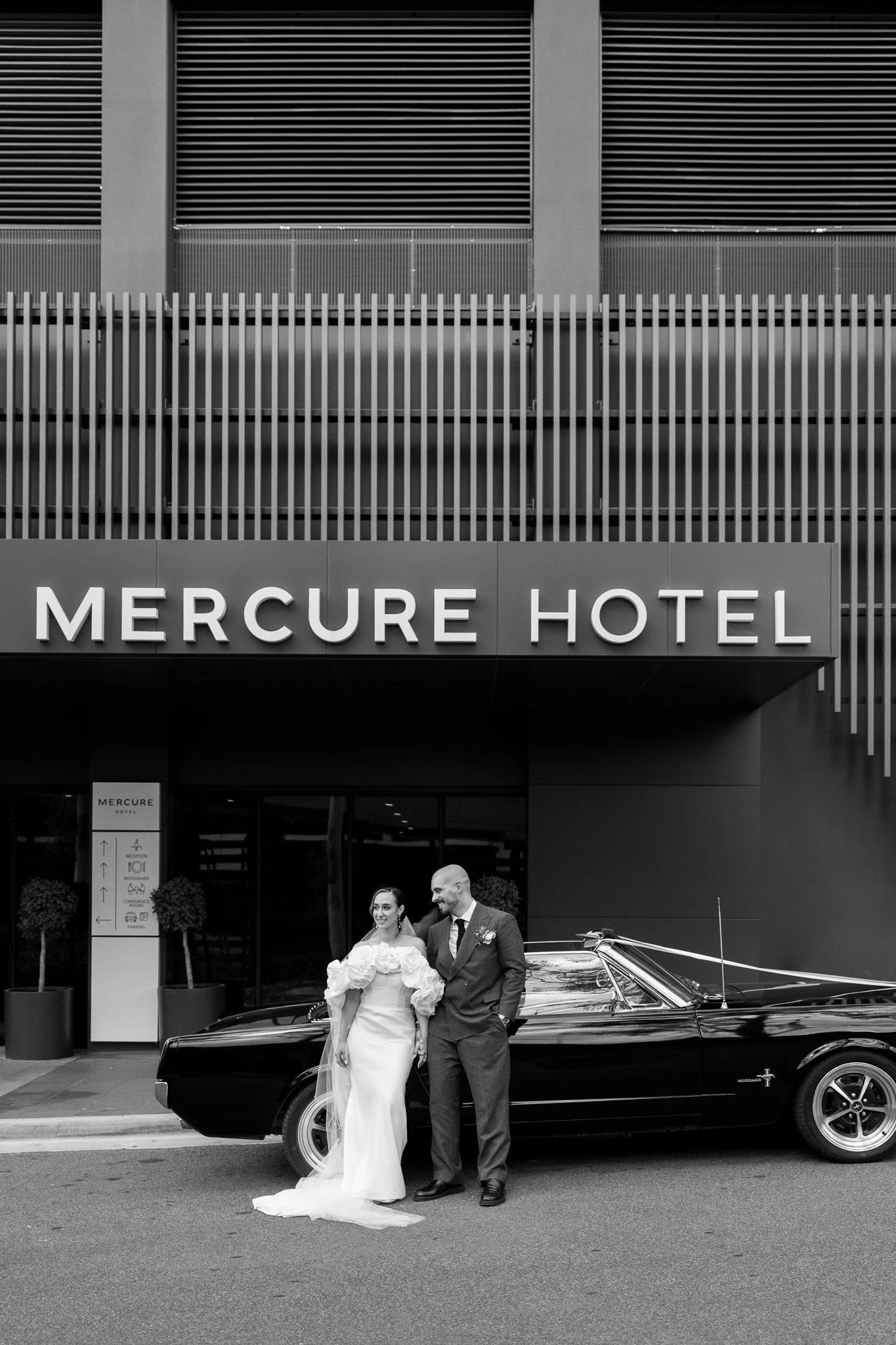 A bride and groom in wedding attire standing in front of a black vintage car outside the Mercure Hotel.