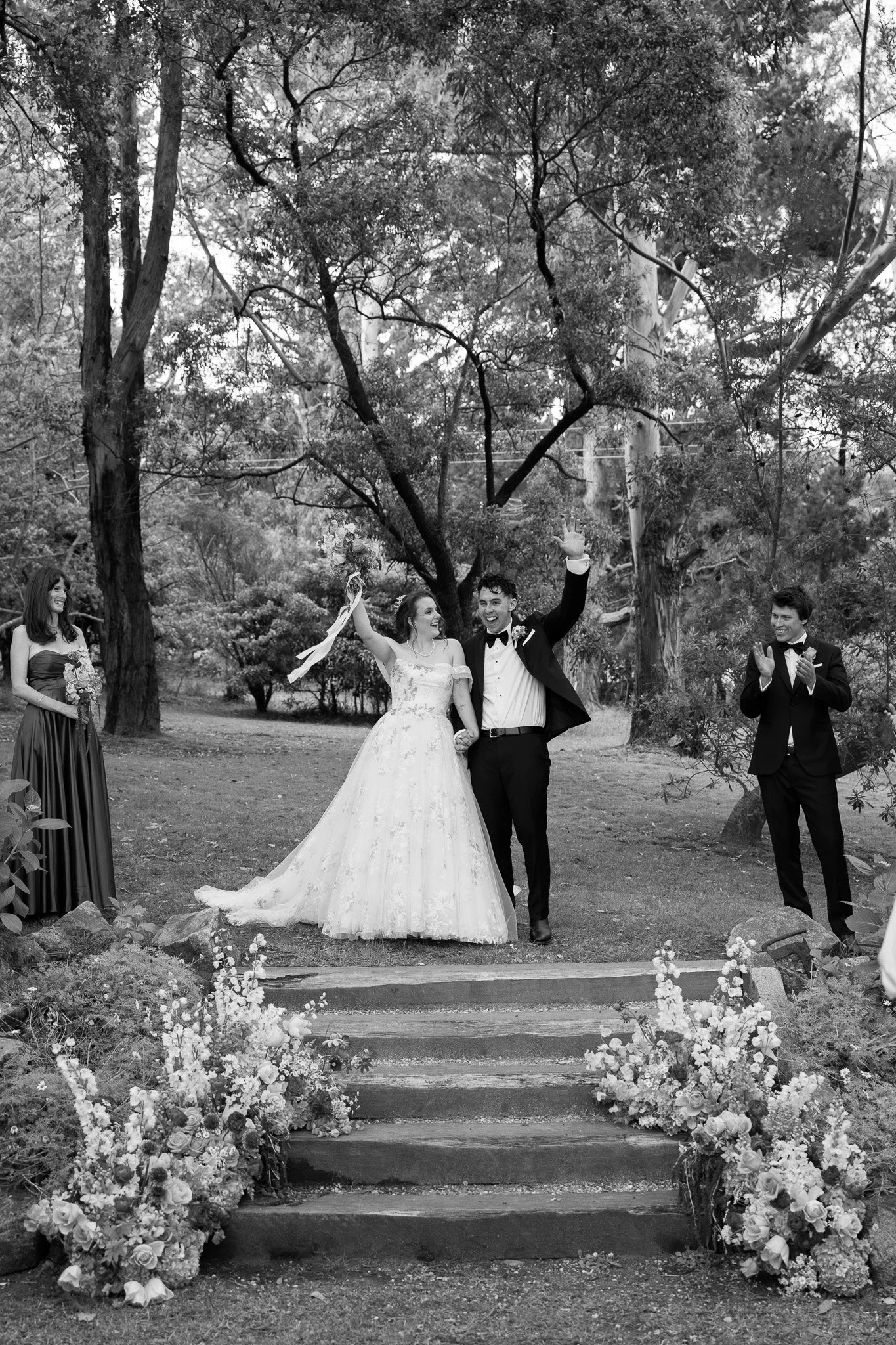 A black and white photo of a wedding ceremony outdoors in a wooded park. The bride and groom are holding hands and smiling as they walk down a small set of steps, with the bride holding a bouquet and the groom waving. Two attendants stand on either s