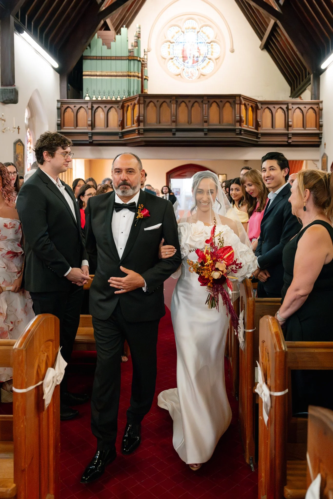 A bride in a white wedding gown and veil walking down the aisle with an older man in a black tuxedo, surrounded by guests inside a church with wooden beams and an organ in the background.