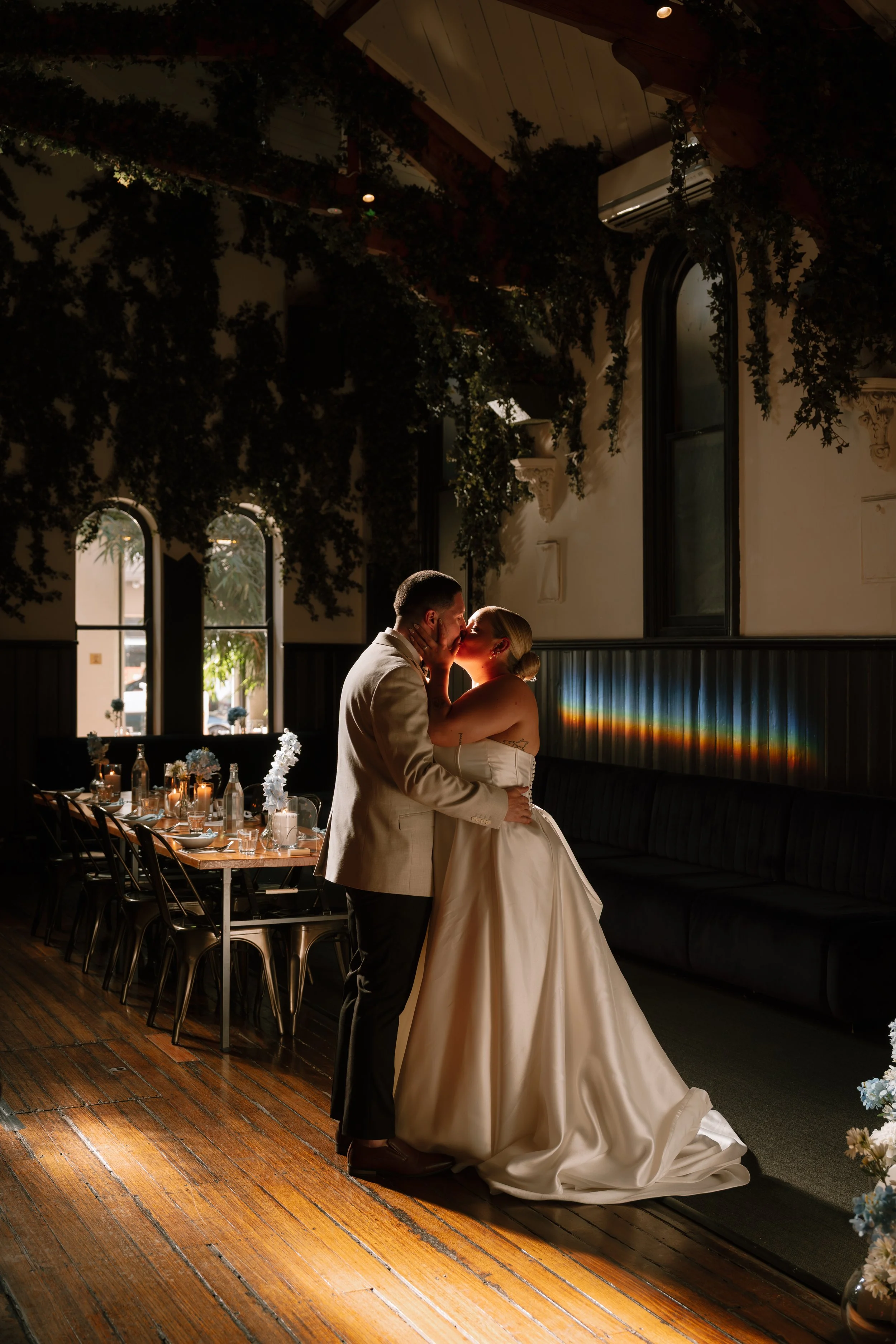 A bride and groom share a kiss during their wedding reception indoors. The bride wears a white gown and the groom a beige suit. The room has dark walls, arched windows, and floral decorations with candles and flowers on a table nearby.