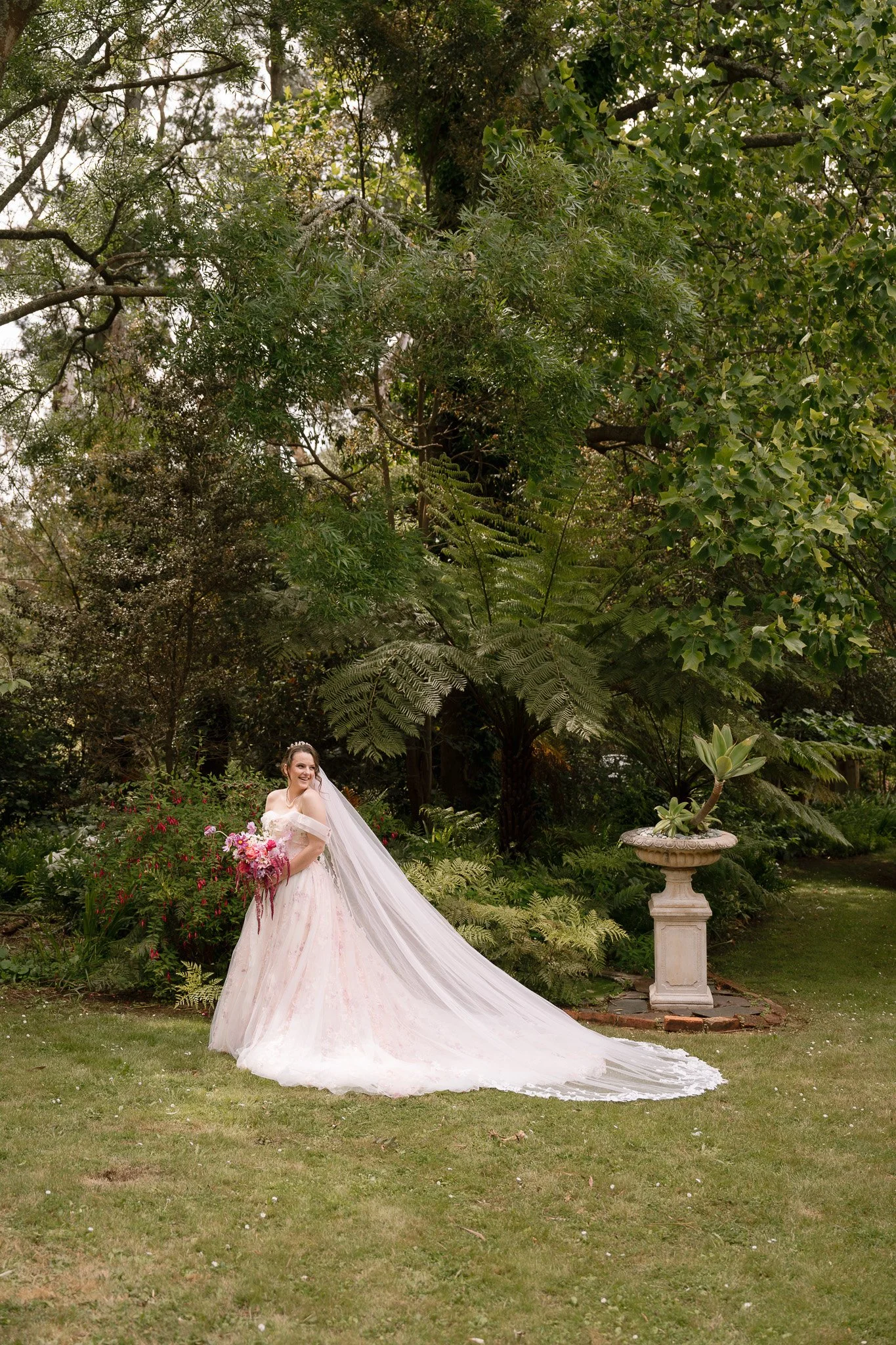 A bride standing outdoors in a garden, holding a bouquet of pink and purple flowers, wearing a long white wedding dress with a veil, smiling amid lush greenery and a stone garden ornament.