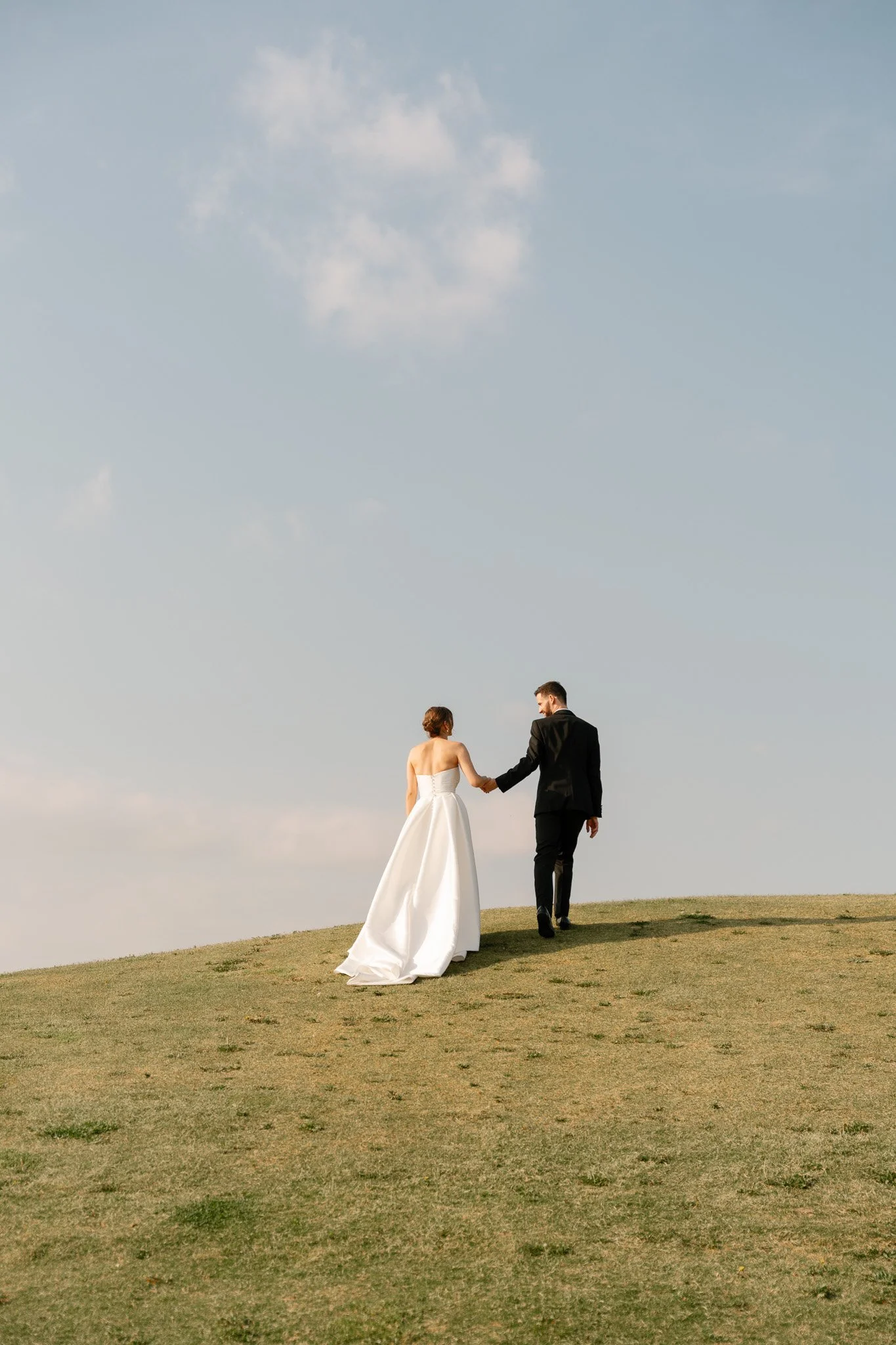 A bride and groom walking hand in hand on a grassy hill, with a clear sky overhead.