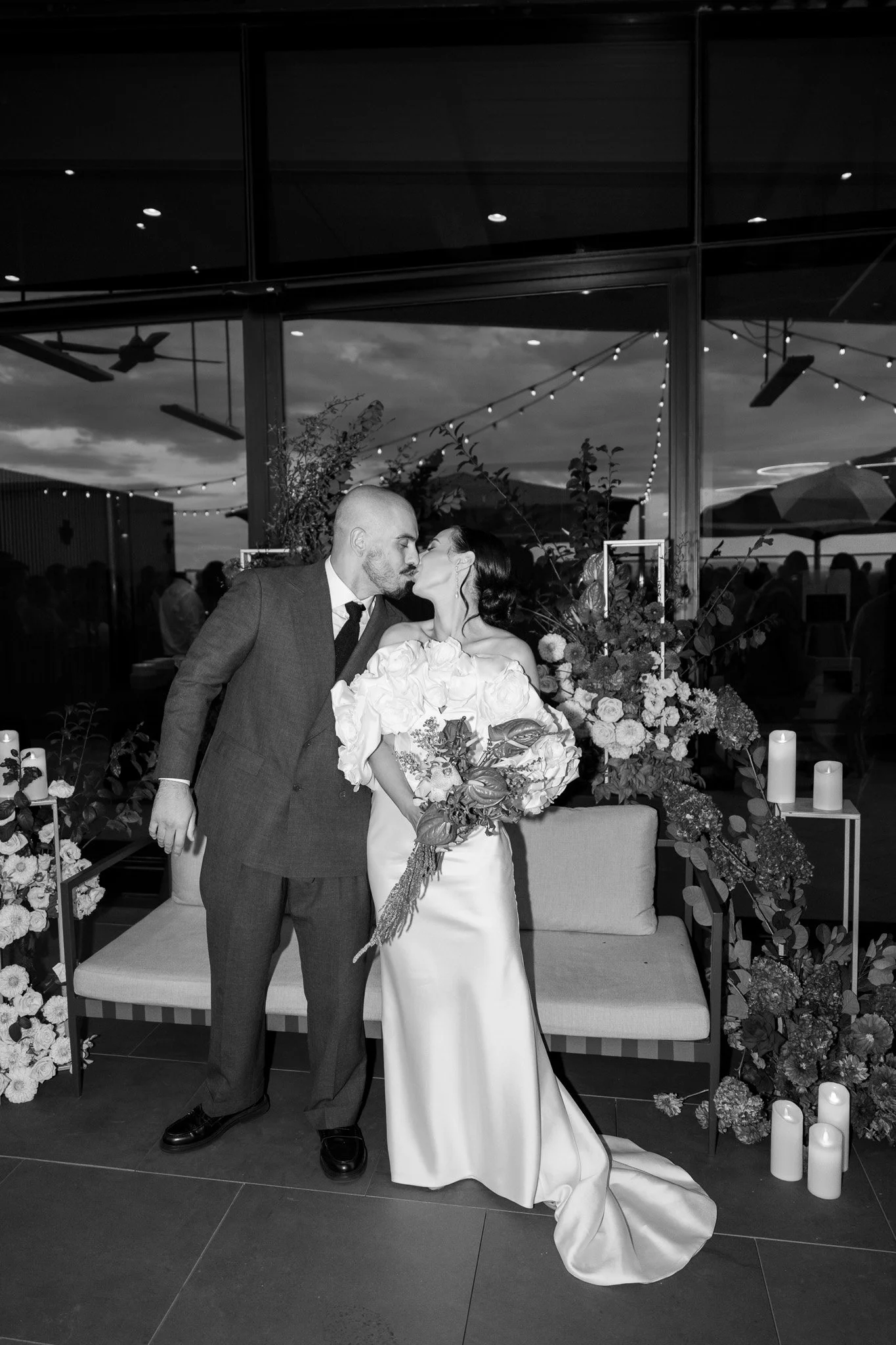 A black and white photo of a newly married couple sharing a kiss, standing in front of floral arrangements and candles, with a large window and outdoor view in the background.