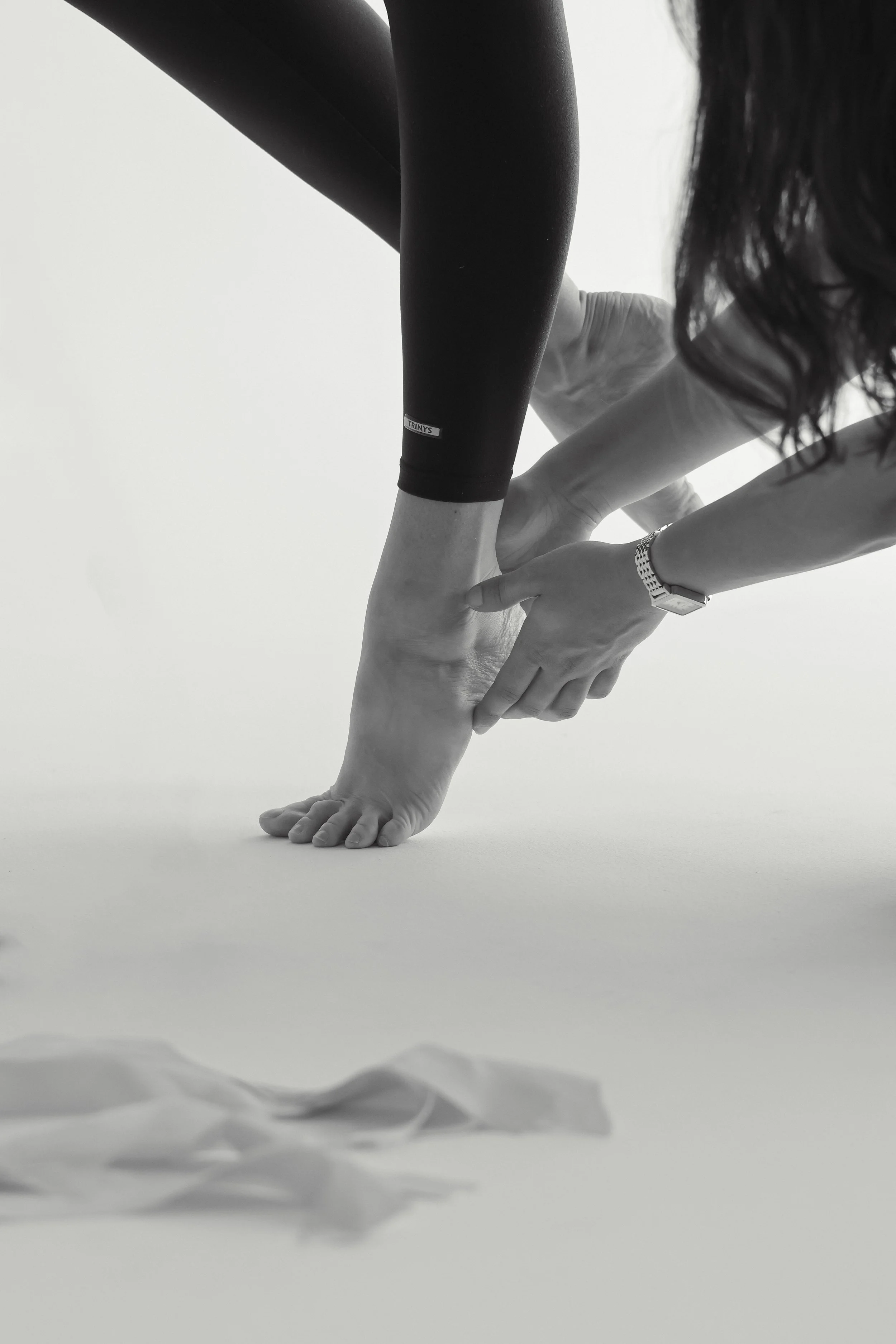 A dance physiotherapist is kneeling on the ground assessing a dancer who is performing a single-leg heel raise. On the bottom left corner of the image there are ribbons from pointe shoes.