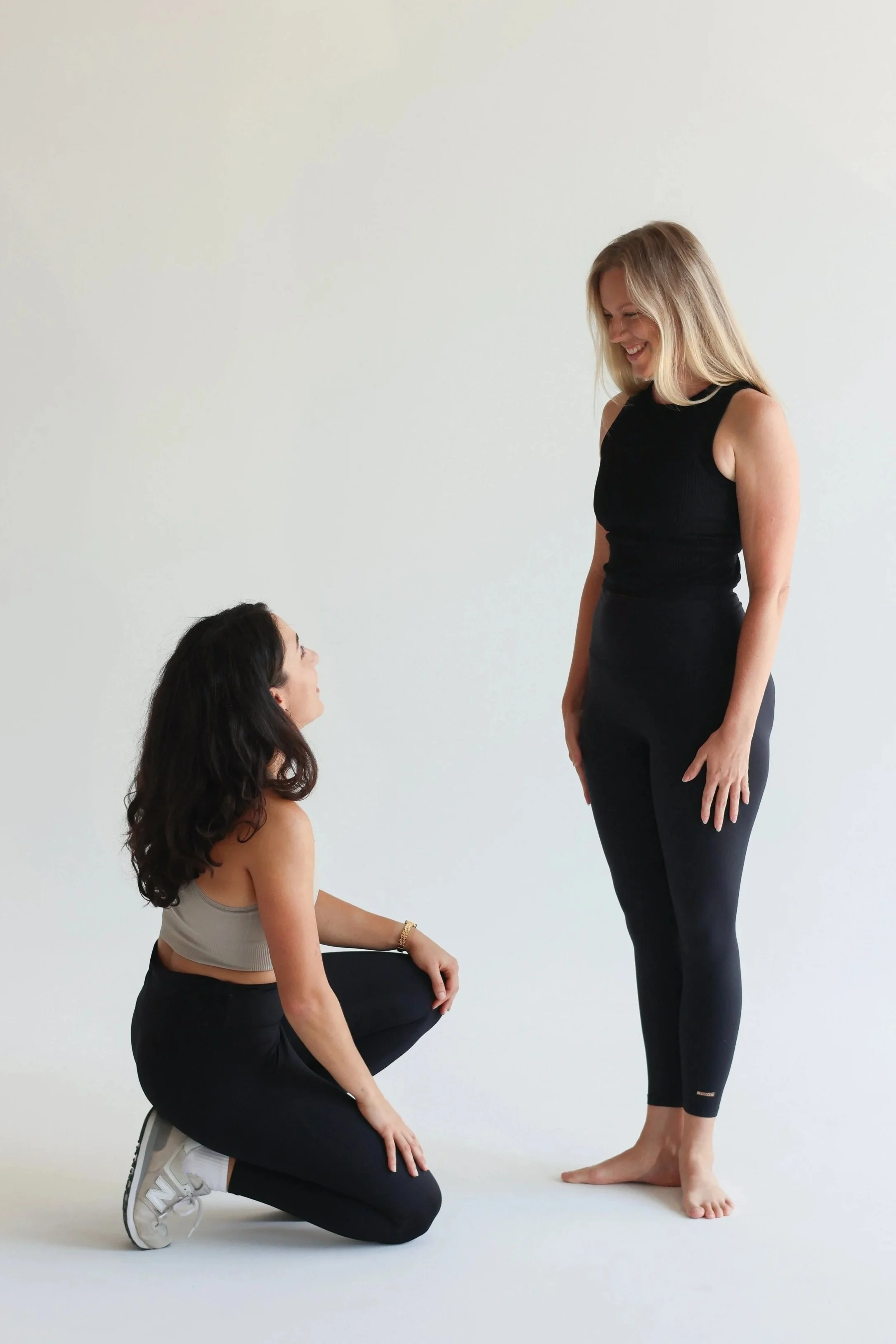 A dance physio is kneeling on the ground looking up at a dancer standing in first position in front of her, they are smiling at each other.