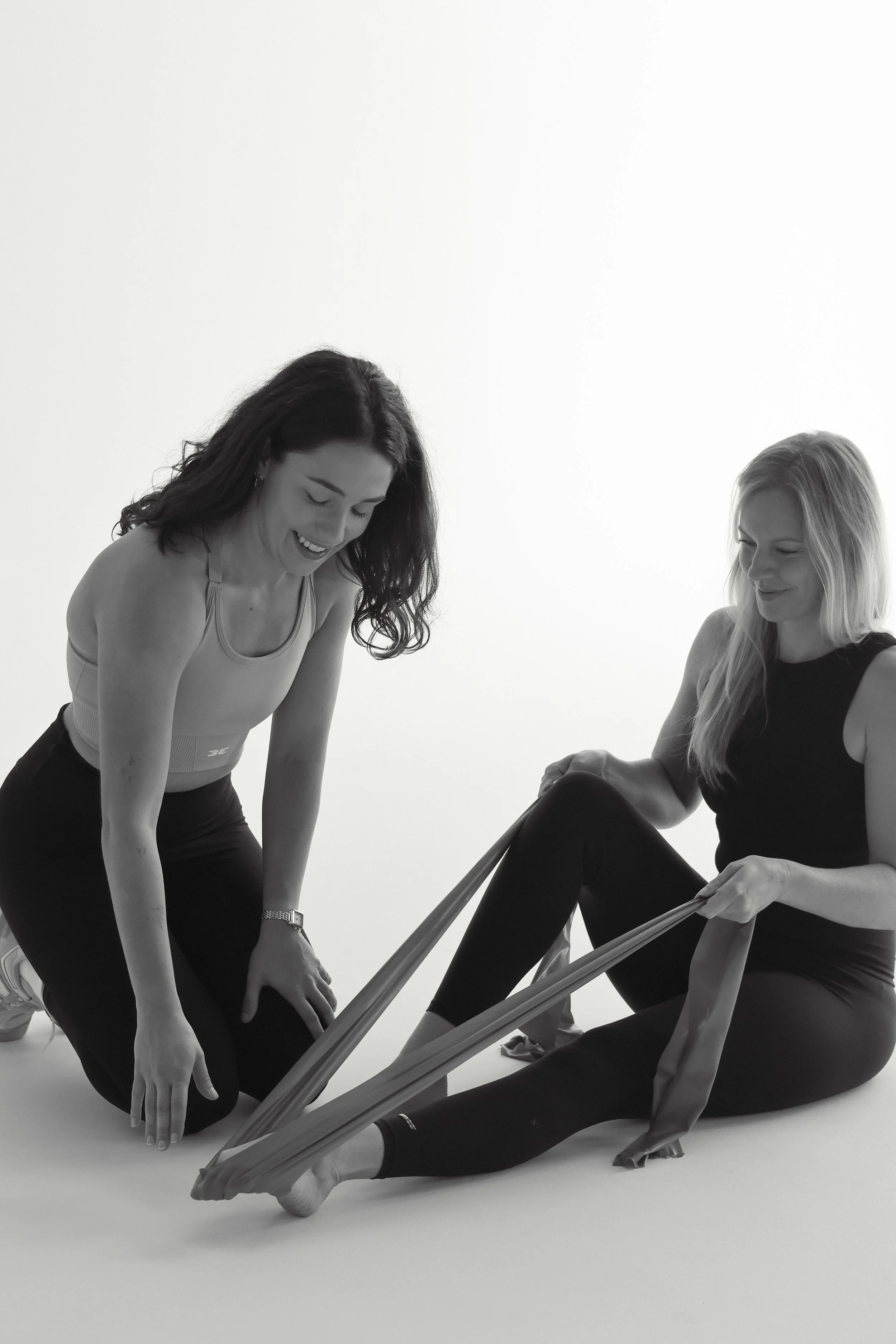 A dance physiotherapist is kneeling on the ground with a dancer in front of her sitting on the floor performing an ankle strengthening exercise with a resistance band. The dance physiotherapist is guiding the dancer through the exercise.