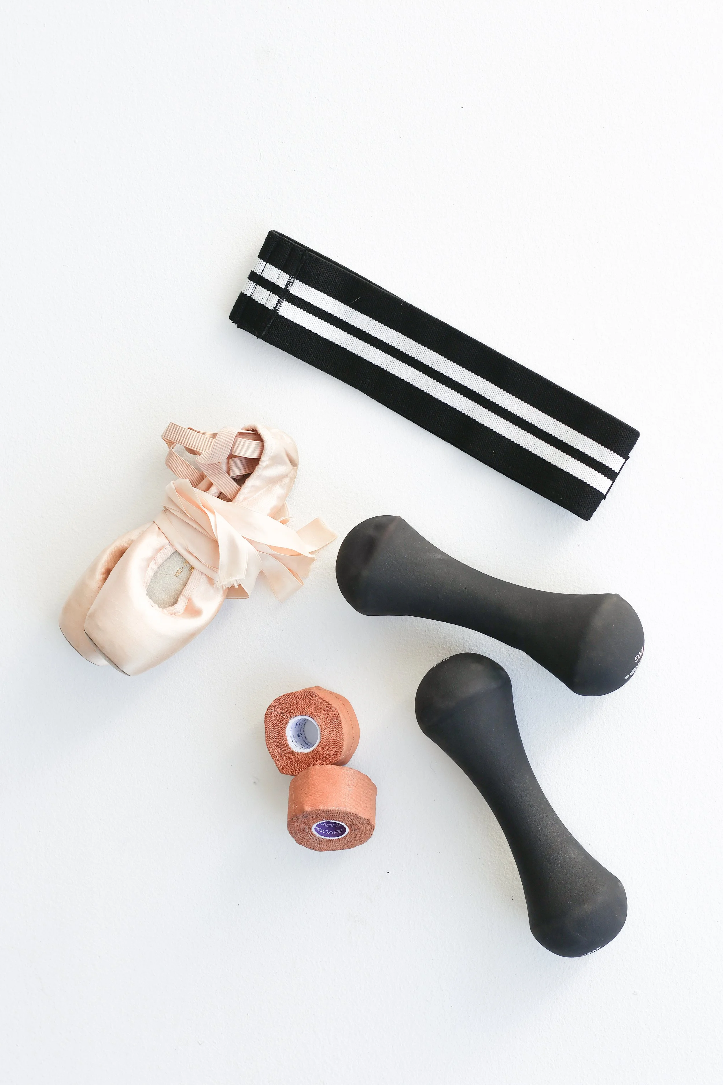 A flat lay of a resistance band, two dumbbells, two rolls of strapping tape, and a pair of pointe shoes used by a dance physiotherapist