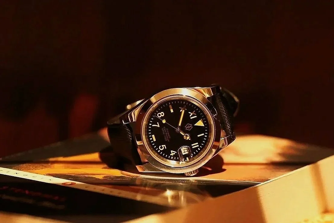 A wristwatch with a black dial, silver bezel, and black strap resting on an open book with warm lighting.