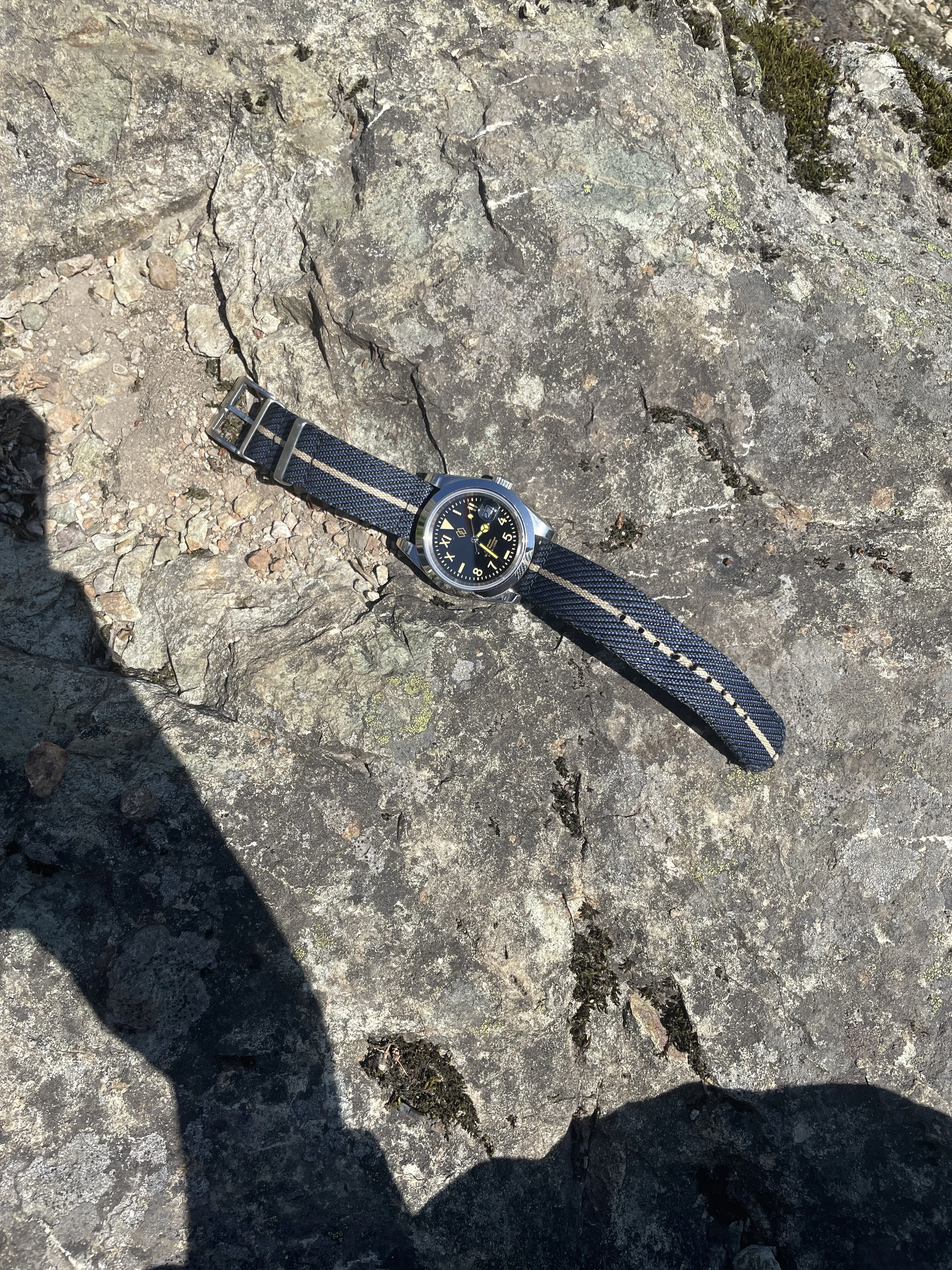 A wristwatch with a black dial and yellow accents placed on a textured rock surface outdoors.