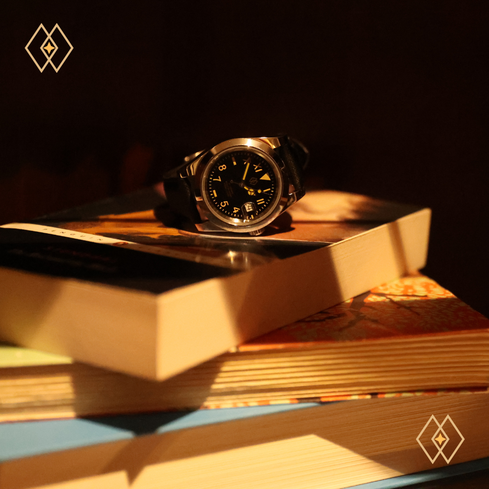 A wristwatch with a dark face and yellow markings on top of stacked books in a dimly lit setting.