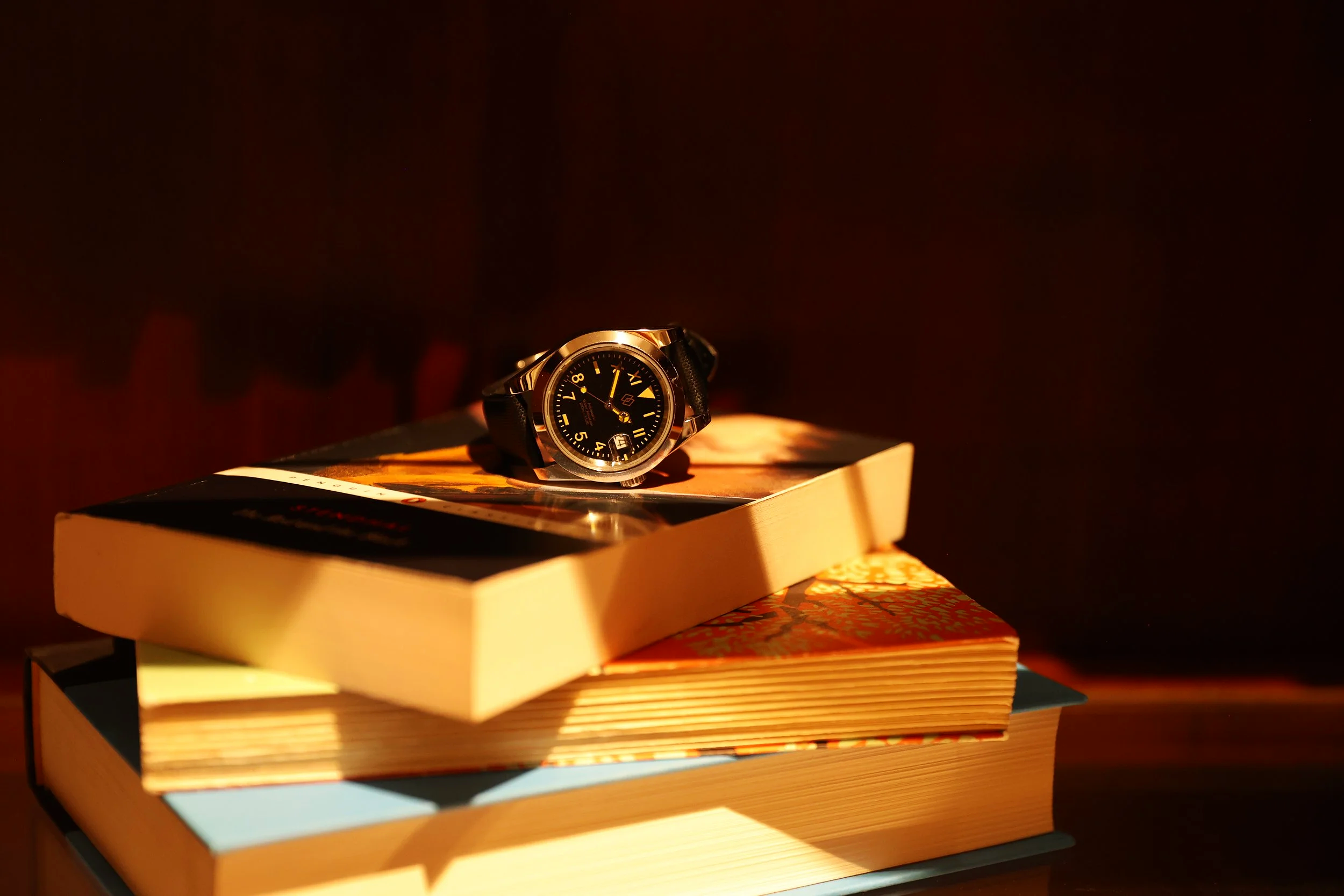 A wristwatch with a black strap and black dial resting on a stack of books with warm lighting.