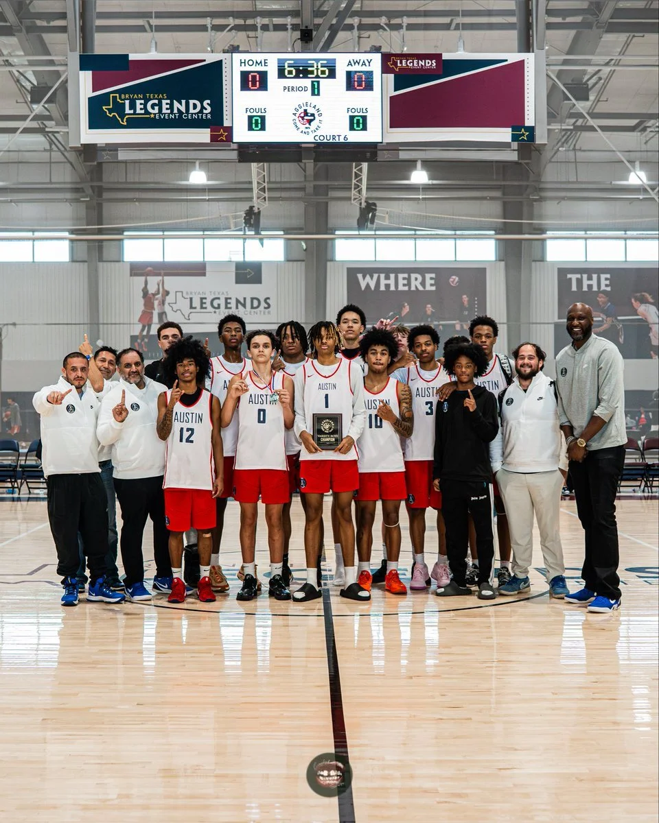 A basketball team in red and white uniforms, along with their coaches, poses for a group photo on the court at Bryan Texas Legends Event Center. The team is holding a championship trophy for the Come and Take It Tip-Off Tournament.