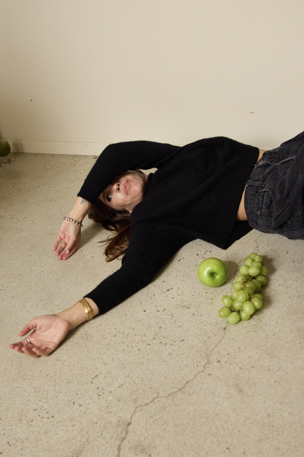 A woman lying on the floor with her arm stretched out, eyeing the camera, with a green apple and a bunch of green grapes next to her.